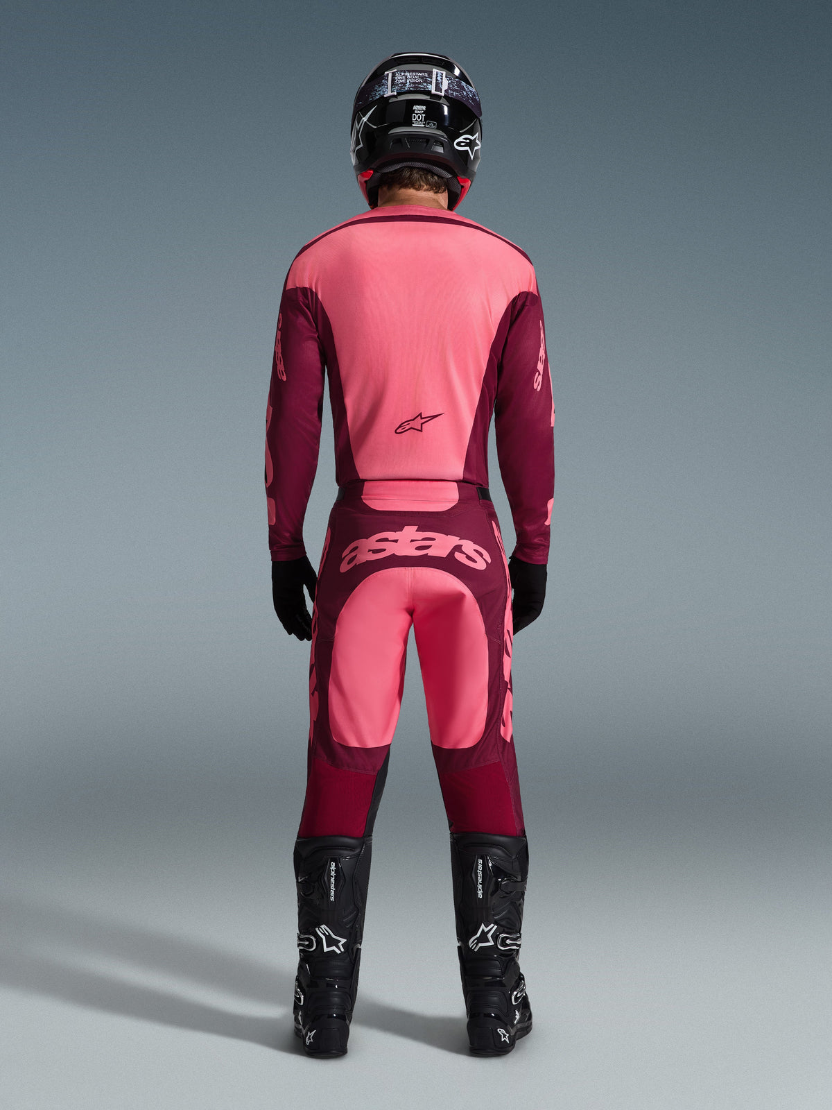 Motocross-Fahrer von hinten im Alpinestars 2026 Racer Riway Trikot und passender Hose in Dark Red Coral, schwarze Stiefel, schwarzer Helm und schwarze Handschuhe, koordinierte Hochleistungs-Ausrüstung für den Offroad-Rennsport