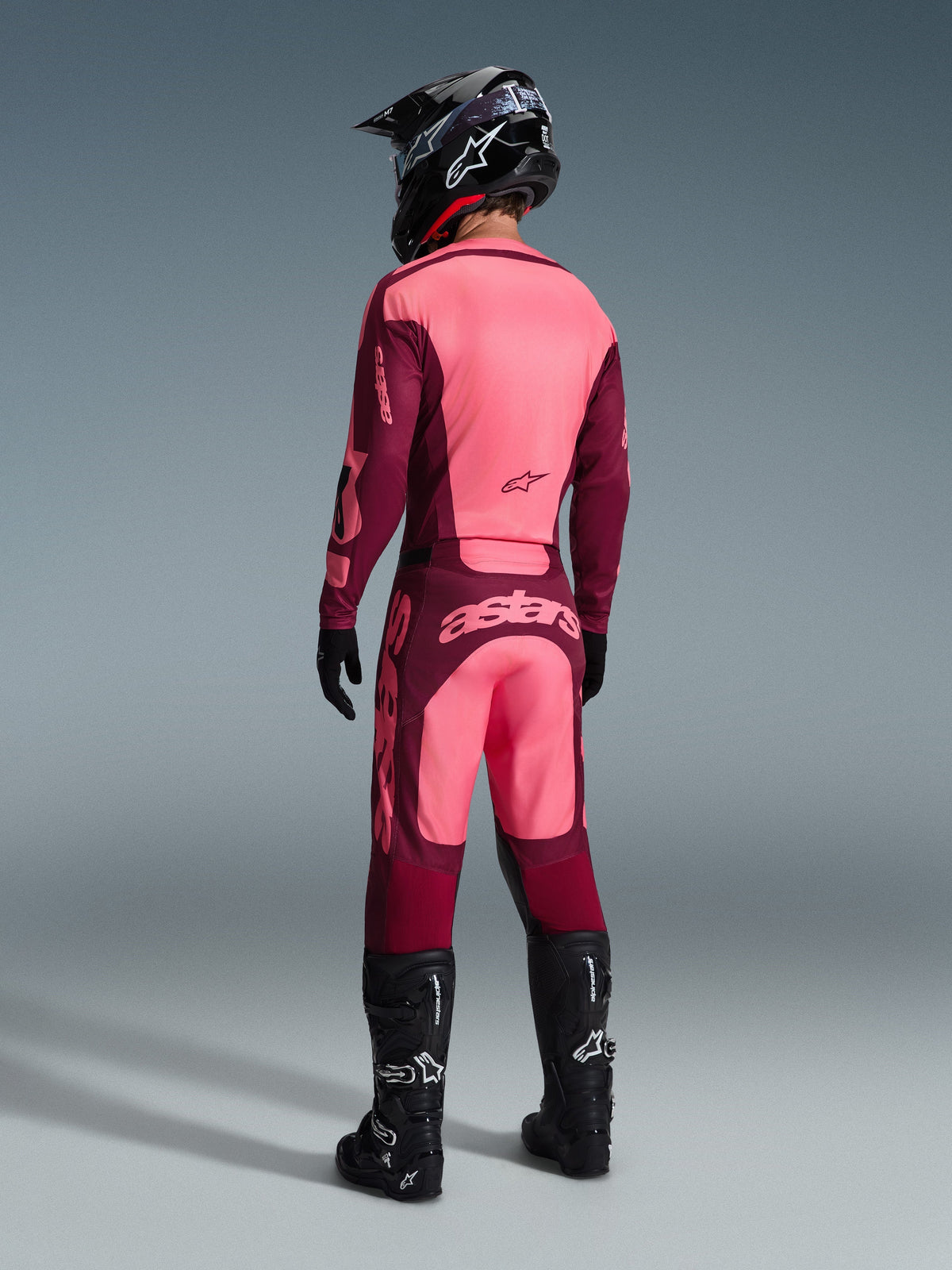 Alpinestars 2026 Racer Riway Trikot und passende Hose, Motocross-Ausrüstung in Dark Red Coral, Rückansicht eines Fahrers mit schwarzem Helm und schwarzen Stiefeln, mit leuchtenden korallenroten und weinroten Panels sowie auffälligem Astars-Branding auf der Rückseite