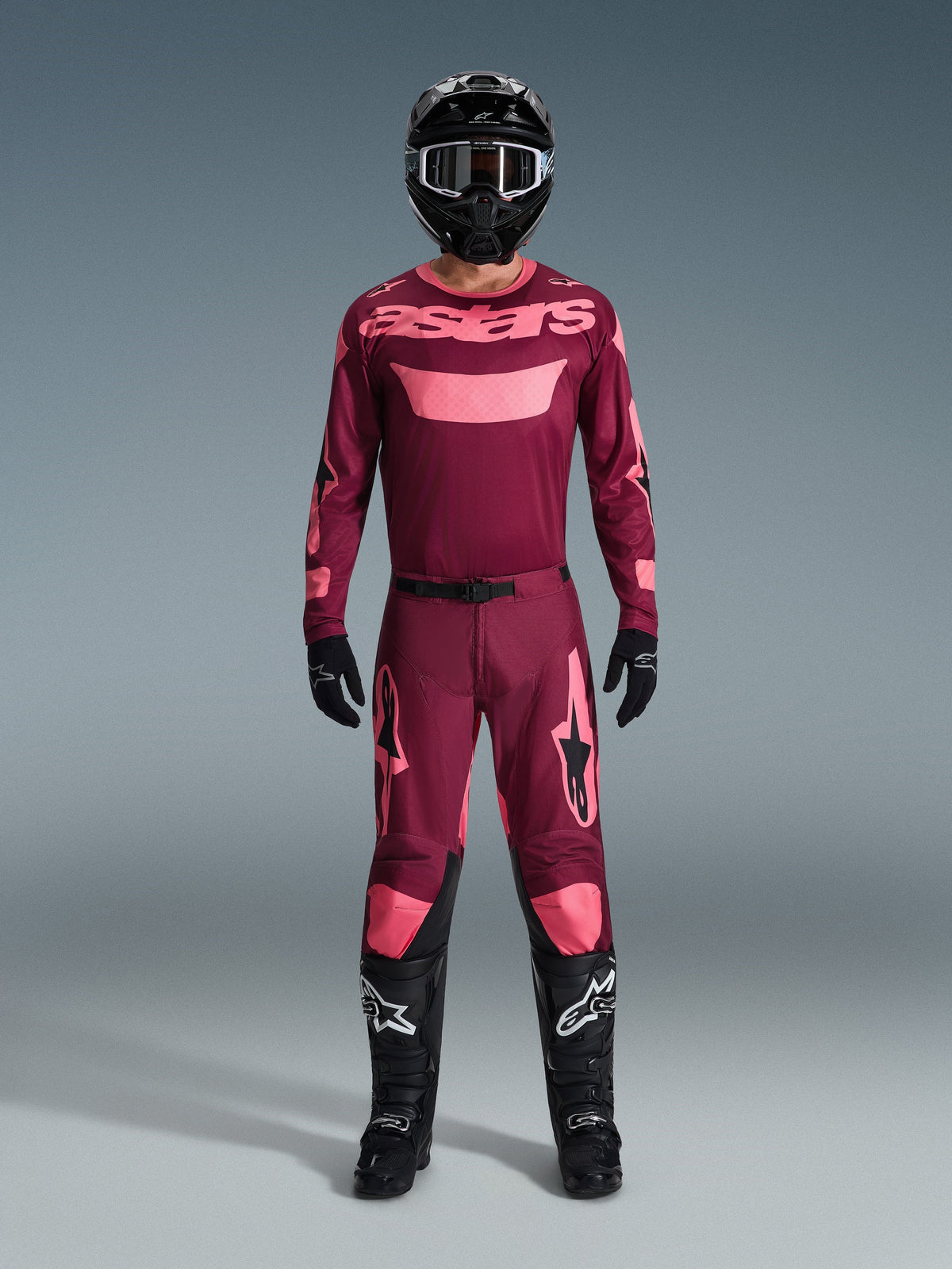 Motocross-Fahrer in kompletter Rennausrüstung, 2026 Racer Riway Trikot in Dark Red Coral, passende dunkelrote Hose mit korallenfarbenen Akzenten, schwarzer Helm mit weißen Akzenten, schwarze Stiefel mit weißen Logos und schwarze Handschuhe, abgestimmtes Outfit für den Offroad-Rennsport