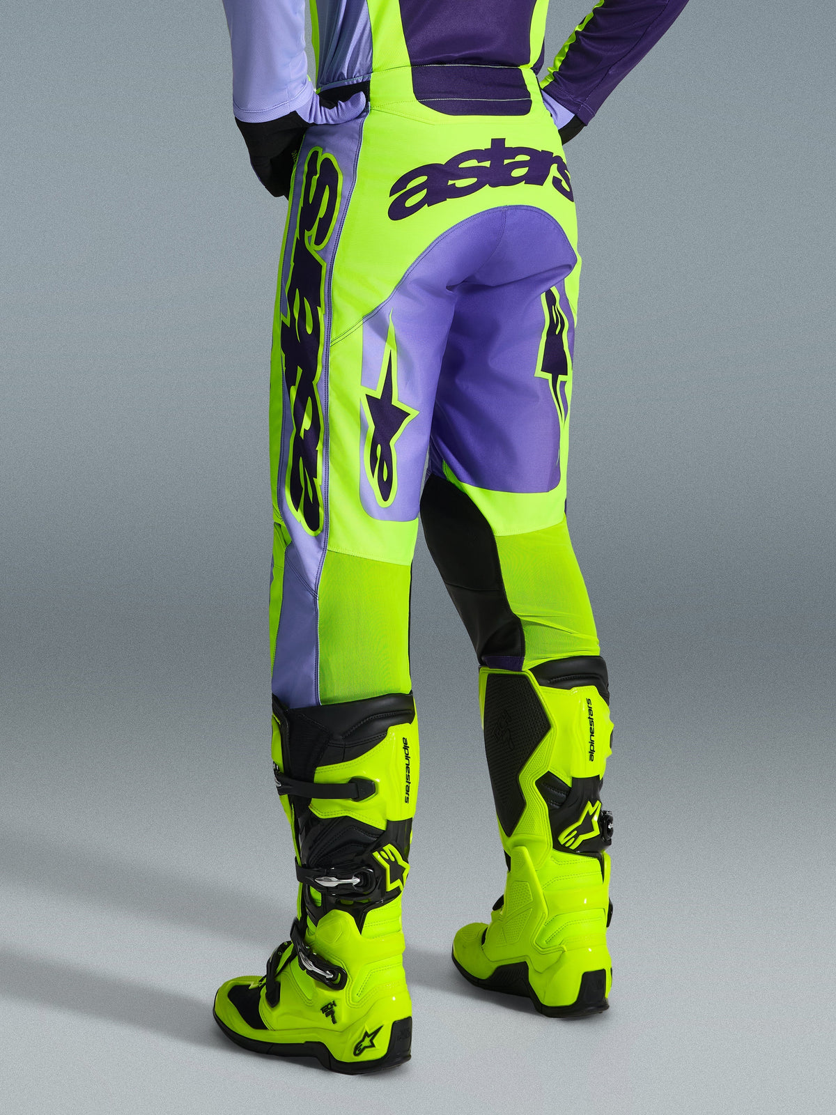Alpinestars 2026 Racer Portl Pants, pantalon de motocross, violet et jaune fluo, vue arrière avec marquage astars imposant et accents violets, porté avec des bottes de motocross jaune fluo assorties, matériau synthétique durable pour la course tout-terrain