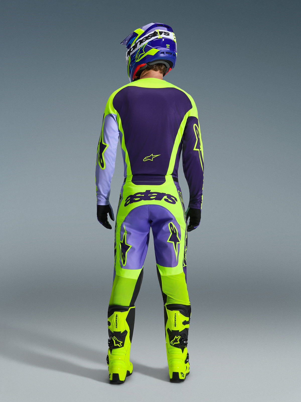 Alpinestars 2026 Racer Portl Pants, pantalon de motocross, violet et jaune fluo, vue arrière complète d'un pilote portant l'équipement complet avec maillot assorti, casque et bottes jaune fluo, avec marquage 'astars' et logos en forme d'étoile.