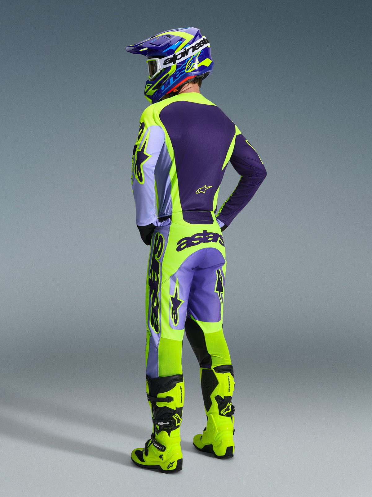 Pilote de motocross vu de dos portant le pantalon Alpinestars Racer Portl 2026 en Purple Yellow Fluo, avec un maillot et un casque violets et jaunes assortis, des bottes de course jaune fluo, logo 'astars' sur l'assise et logos d'étoiles sur les jambes.
