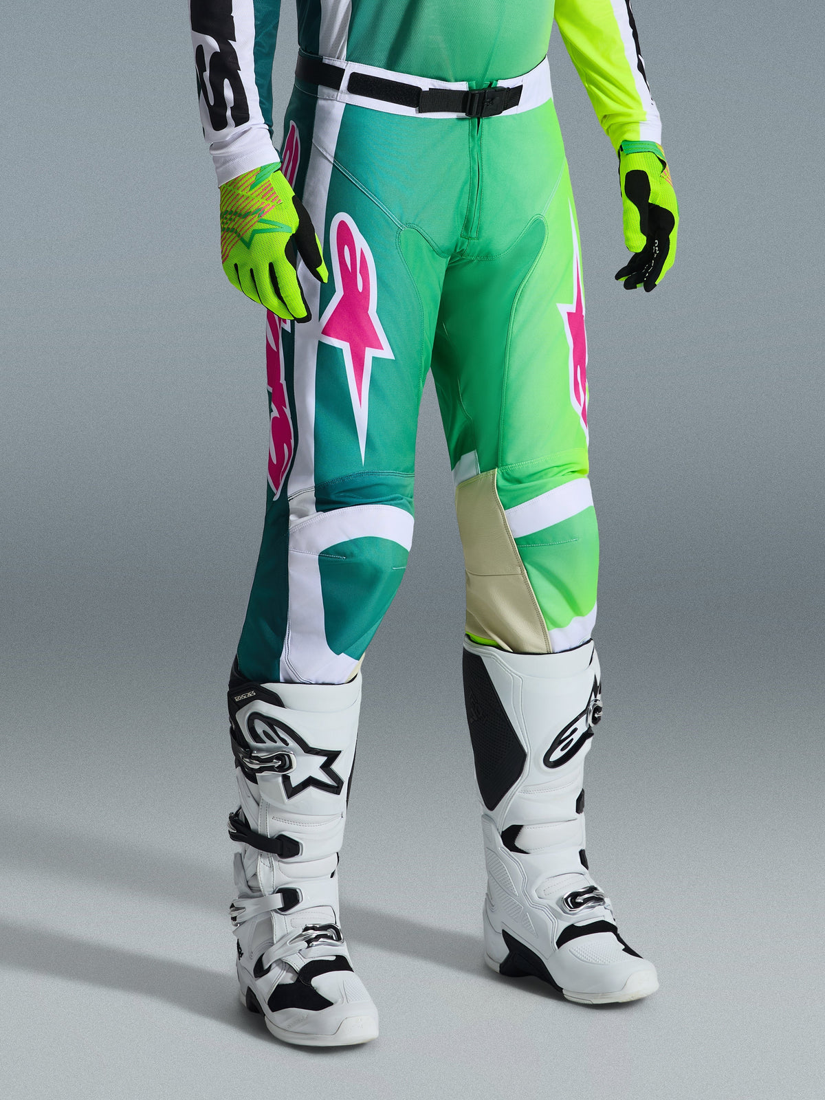 Pantalon Alpinestars 2026 Racer Portl, pantalon de motocross, vert, blanc et noir avec des logos roses, porté avec des bottes blanches et des gants jaune fluo, doté de motifs graphiques audacieux et d'une boucle de taille réglable noire, équipement de performance pour la conduite tout-terrain.