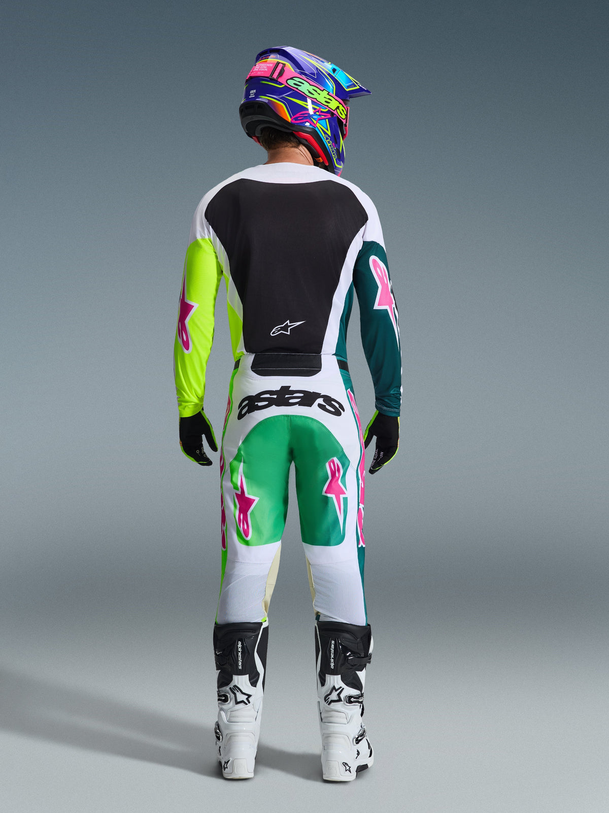Pilote de motocross vu de dos portant le pantalon Alpinestars Racer Portl 2026 vert, blanc et noir, maillot assorti avec des logos d'étoiles roses, casque multicolore « astars » et bottes blanches et noires, ensemble d'équipement de course tout-terrain coordonné.