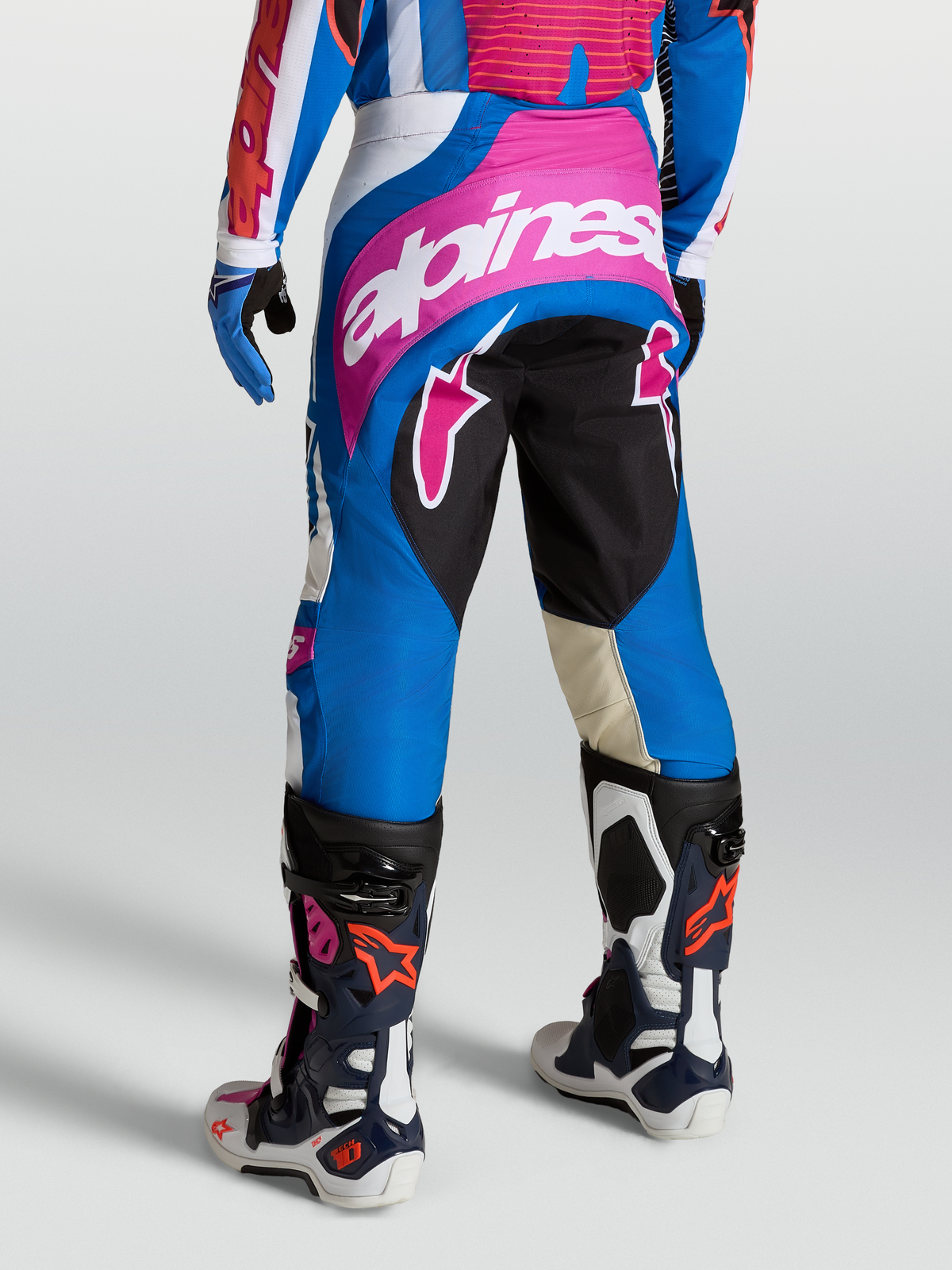 Alpinestars Pantalon Techstar Maven, pantalon de motocross bleu et rose porté avec des bottes Tech 10, panneaux extensibles et logo blanc, profil arrière pour performance de course.