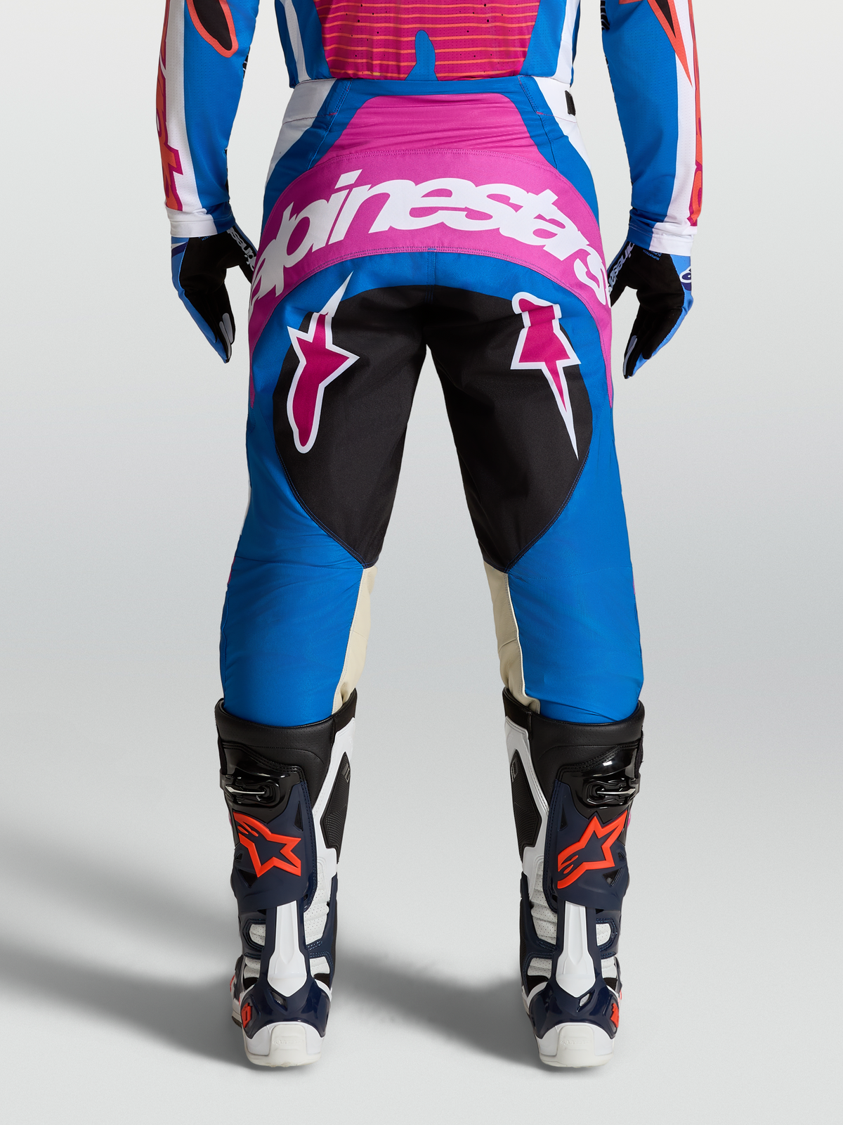 Pantalon Techstar Maven Alpinestars, pantalon de motocross, bleu et rose avec logos blancs, empiècements en cuir de chèvre aux genoux, porté avec des bottes de moto.