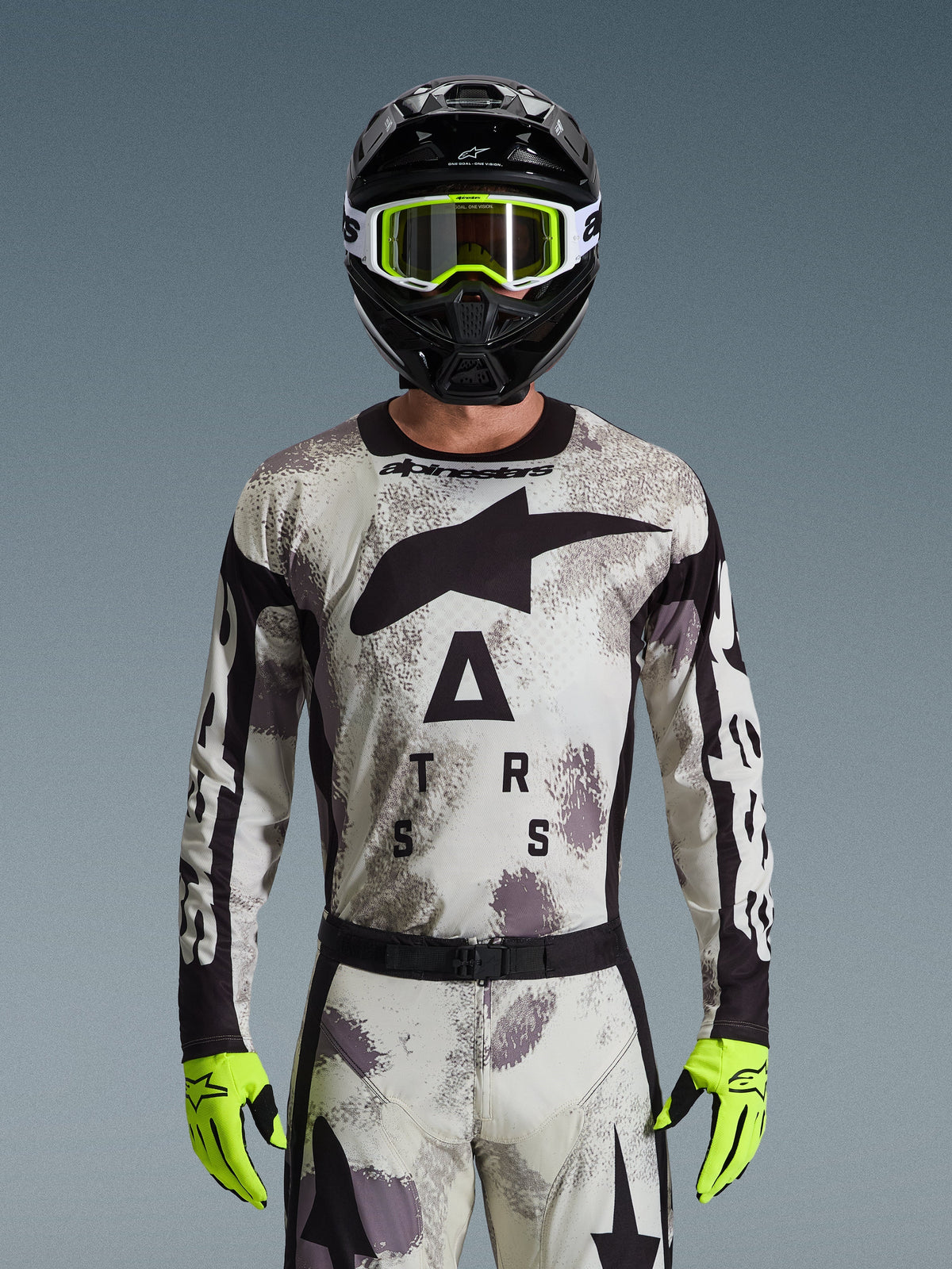 Maillot Alpinestars Racer Lahnd 2026, ensemble d'équipement de motocross, motif Sand Camo gris et blanc, grand logo étoile noire et lettrage « ASTARS », porté avec un casque noir, des lunettes jaune fluo et des gants jaune fluo, équipement professionnel de course tout-terrain