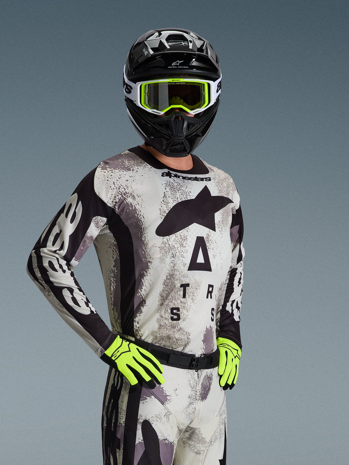 Pilote de motocross portant le maillot et l'ensemble d'équipement Alpinestars Racer Lahnd 2026 en Sand Camo, motif camouflage gris clair avec logos noirs, casque noir avec lunettes blanches et jaune néon, gants jaune néon éclatant, tenue de course tout-terrain professionnelle