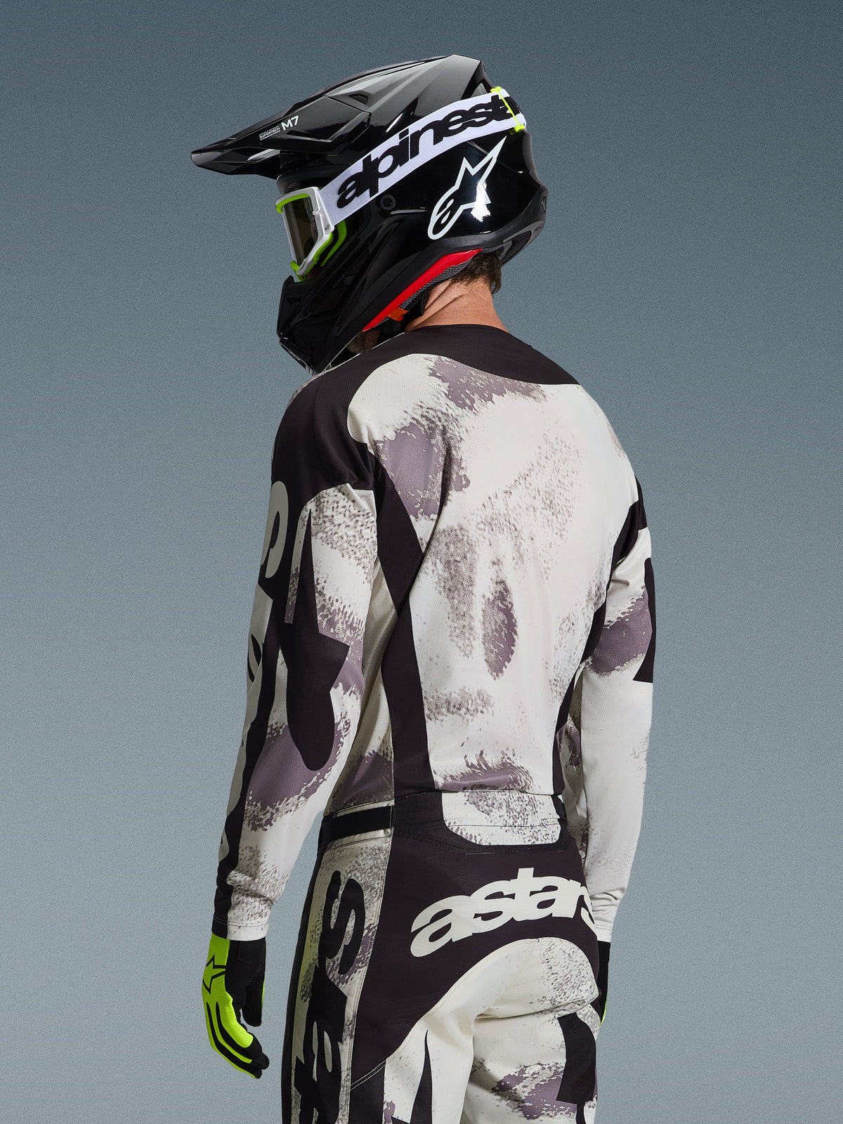 Pilote de motocross de dos portant l'ensemble d'équipement Alpinestars Racer Lahnd 2026, maillot et pantalon Sand Camo gris et blanc avec détails noirs, casque noir avec logo Alpinestars blanc, gants jaune fluo et noirs, tenue de performance tout-terrain coordonnée