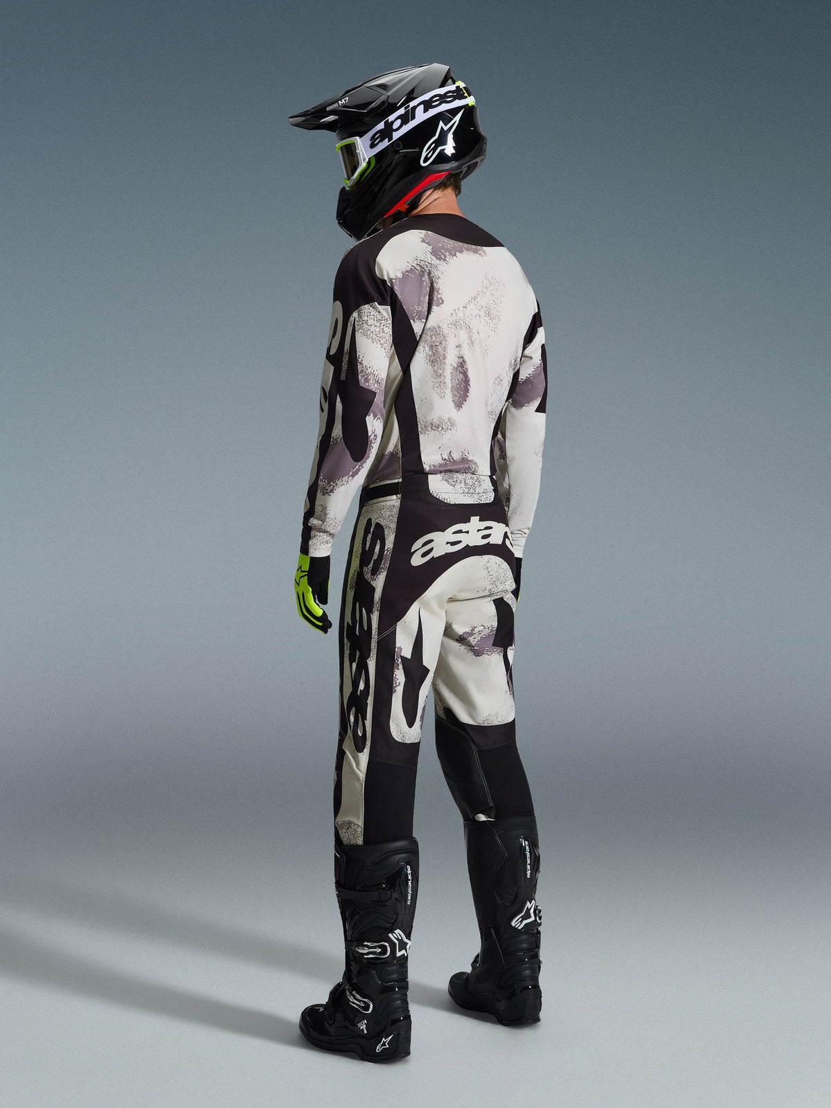 Motorcrossrijder die de Alpinestars 2026 Racer Lahnd-broek in Sand Camo en grijs draagt, volledige race-uitrusting met bijpassend shirt, zwarte helm en zwarte laarzen, achteraanzicht met focus op de opvallende branding en het ergonomische ontwerp.