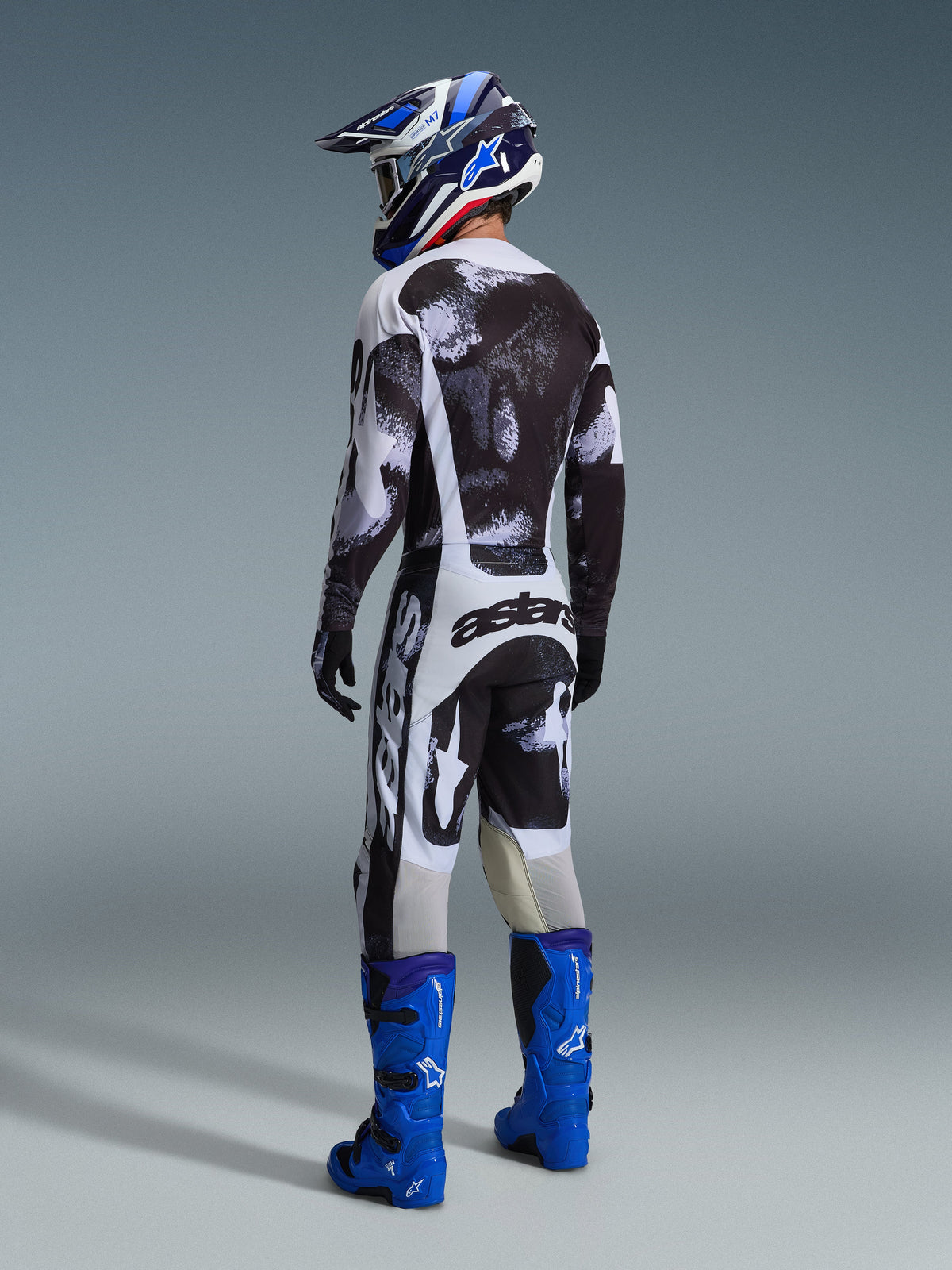 Pilote de motocross portant une tenue de course complète vue de dos, maillot et pantalon Racer Lahnd 2026 en Iron Camo, motif abstrait noir, gris et blanc, bottes Alpinestars bleu vif, casque bleu et blanc, tenue coordonnée pour la course tout-terrain