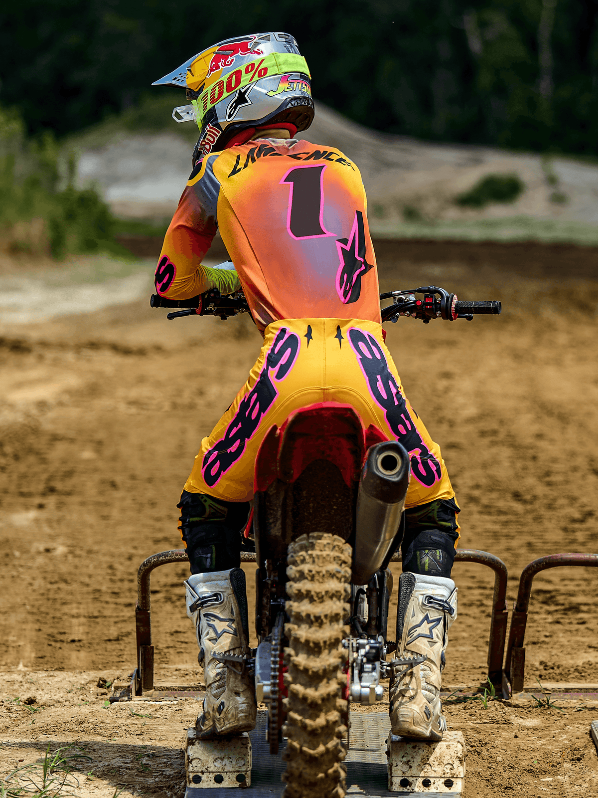 Motocrossrijder van achteren gezien bij een starthek, gekleed in Alpinestars 2025 Supertech Lipan-broek in geel en roze, bijpassende jersey met roze accenten en nummer 1, witte Alpinestars-laarzen en een racehelm, gecoördineerde professionele off-road race-uitrusting.