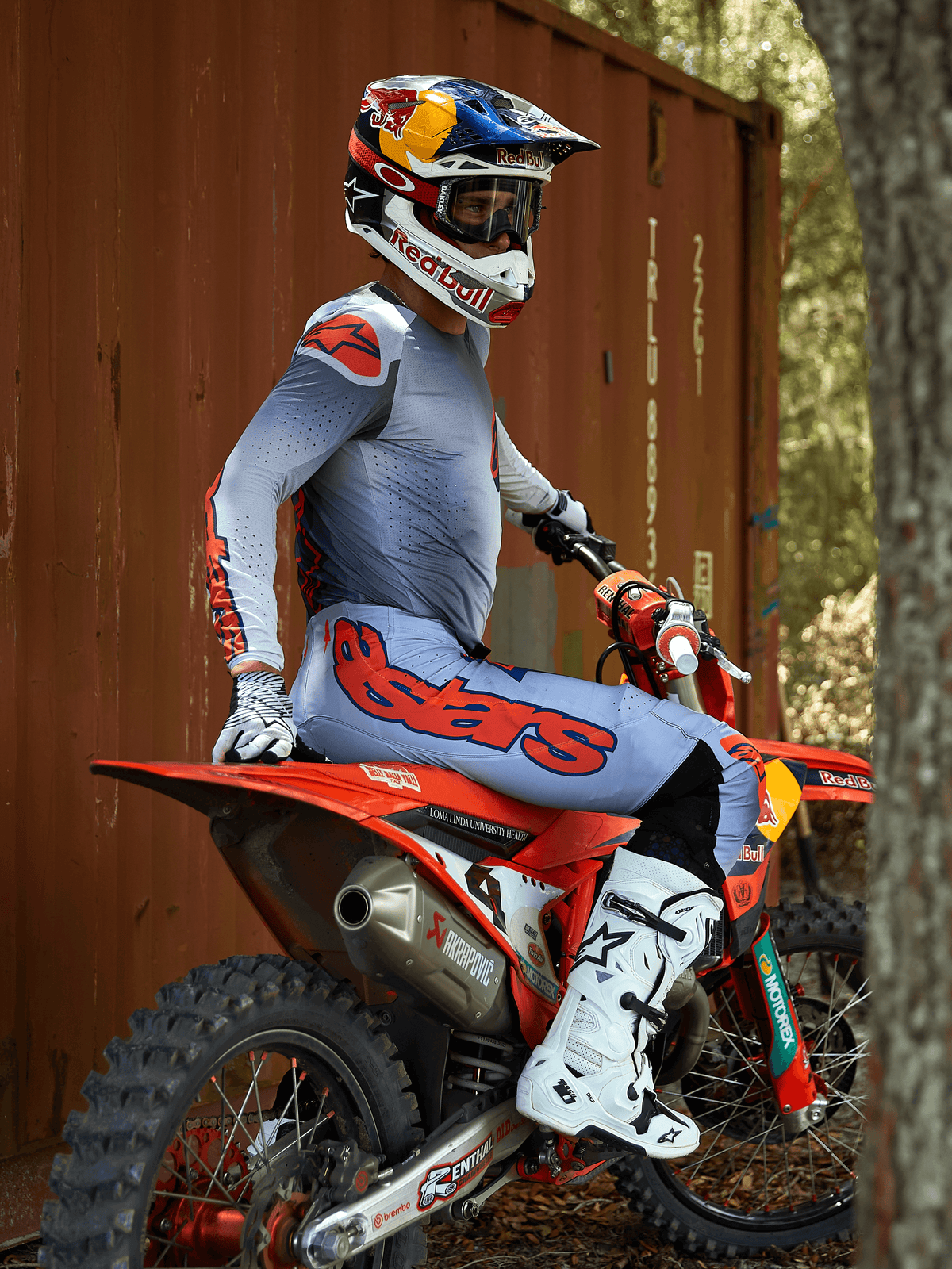 Motocross-Fahrer auf einem roten Dirtbike, trägt das Alpinestars 2025 Supertech Lipan Ausrüstungsset in Grau und Orange Fluo, hellblaue Hose mit markantem orangefarbenem Astars-Logo, passendes Trikot, weiße Motocross-Stiefel und einen Red Bull Helm, professionell abgestimmte Rennausrüstung