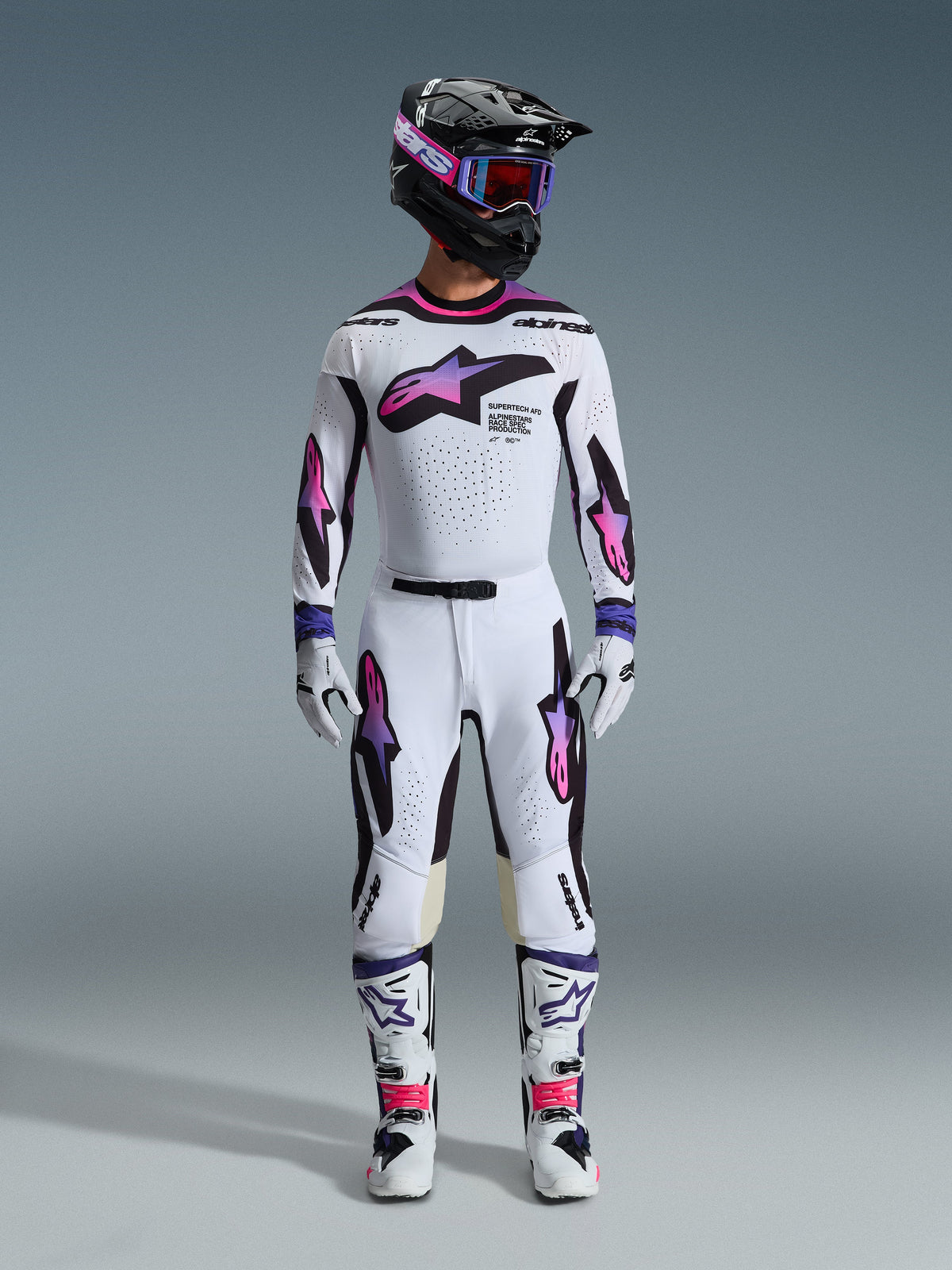 Motocross rider wearing a complete racing gear set, Alpinestars 2026 Supertech AFD Vista Pants and jersey in White Purple and Black, matching gloves, white and black boots, black helmet with purple goggles, sleek athletic fit for off-road performance