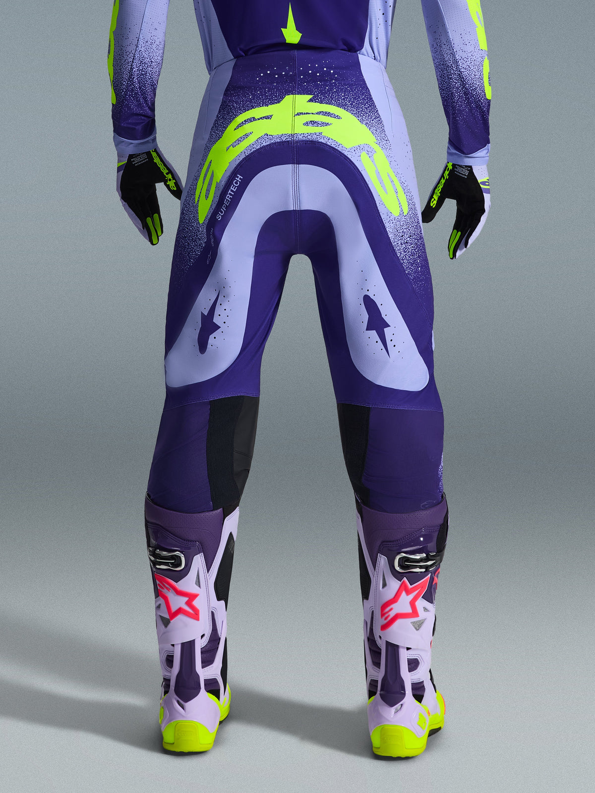 Alpinestars 2026 Supertech Scenz broek, motorcrossbroek, paars en fluogeel, achteraanzicht met groot geel logo op het zitvlak, gedragen met bijpassende jersey, handschoenen en beschermende laarzen, atletische pasvorm voor professionele motorcrossraces