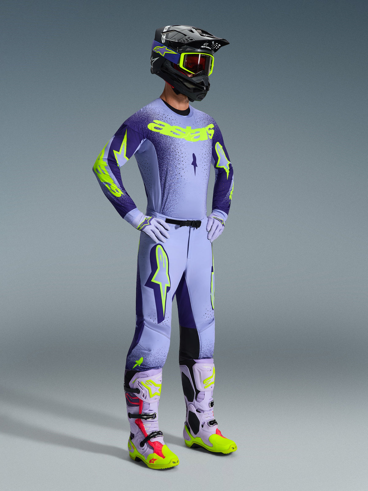 Alpinestars 2026 Supertech Scenz broek, motorcrossbroek, paars en geel fluo, gedragen door een rijder in een volledige race-outfit met bijpassende jersey met kleurverloop, lila handschoenen en bijbehorende geel-paarse laarzen, voorzien van een zwarte helm en bril.