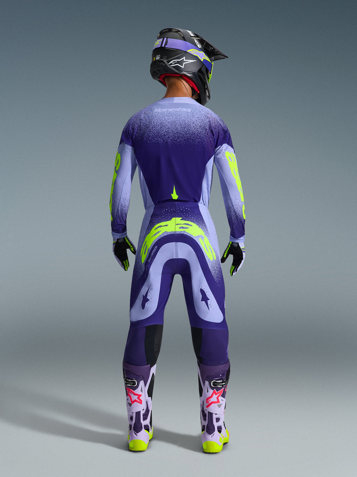 Motorcrosser van achteren gezien in Alpinestars 2026 Supertech Scenz broek in Purple Yellow Fluo, bijpassende jersey met paars verloop en neongele graphics, zwarte helm en paarse laarzen, gecoördineerde professionele race-uitrusting voor off-road prestaties.