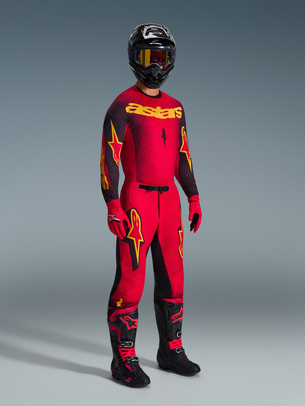 Alpinestars 2026 Supertech Scenz Pants, pantalon de motocross, rouge, noir et jaune, porté par un pilote avec une tenue de course complète comprenant un maillot et des bottes assortis, avec des logos d'étoiles jaunes et noirs et une coupe athlétique