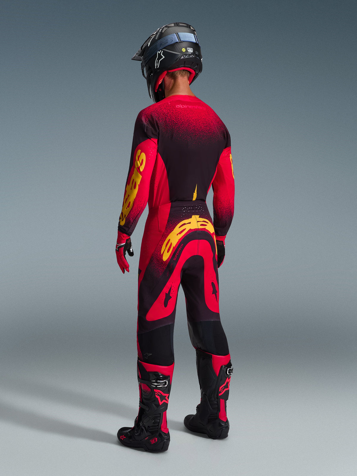 Pilote de motocross vu de dos portant le pantalon Alpinestars Supertech Scenz 2026 en Rouge Noir Jaune, maillot assorti avec logo jaune vif, bottes de motocross noires et rouges, casque noir, ensemble complet d'équipement de course professionnel pour la performance tout-terrain.