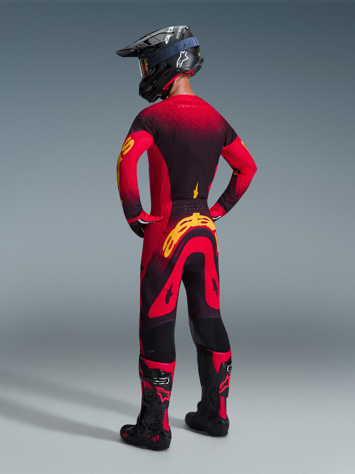 Pilote de motocross de dos portant le pantalon Alpinestars Supertech Scenz 2026 en rouge, noir et jaune, avec un grand logo jaune sur le fessier, maillot dégradé rouge et noir assorti, casque noir et bottes de motocross rouges, équipement de course professionnel pour la performance tout-terrain