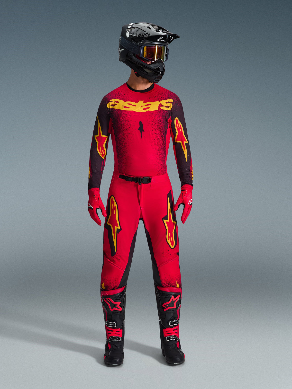 Pantalon Alpinestars Supertech Scenz 2026, pantalon de motocross, rouge, noir et jaune, porté par un pilote avec une tenue de course complète, maillot assorti rouge et noir avec logos jaunes, casque noir, gants rouges et bottes noires, design athlétique pro-fit pour la compétition tout-terrain