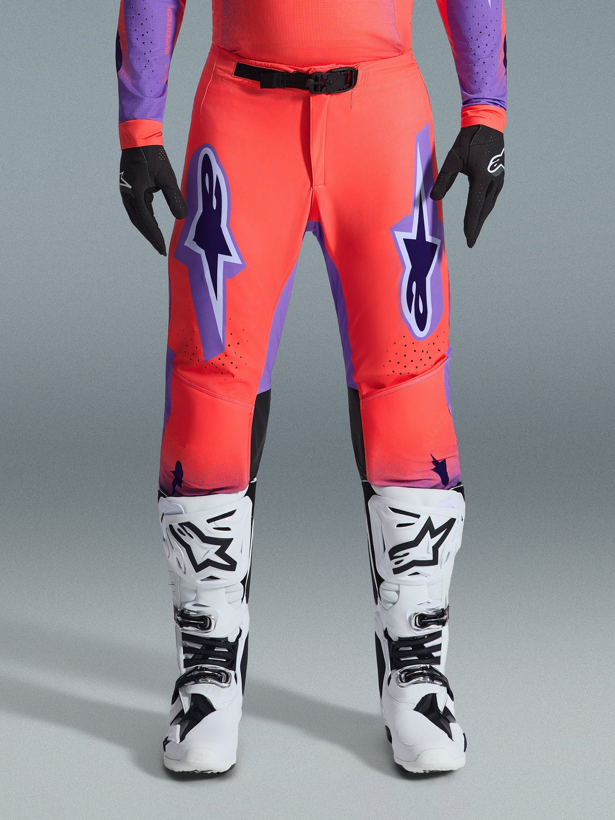 Alpinestars 2026 Supertech Scenz crossbroek, motocrossbroek, Hot Coral en paars, gedragen met bijpassende jersey, wit-zwarte laarzen en zwarte handschoenen, voorzien van grote paarse Astars-logo's op de dijen en een zwarte magnetische gesp, professionele atletische racepasvorm