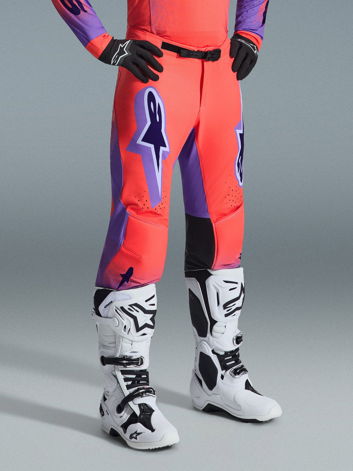 Alpinestars Supertech Scenz broek 2026, motorcrossbroek, Hot Coral en paars, gedragen met wit-zwarte motorcrosslaarzen en zwarte handschoenen, voorzien van een groot paars Alpinestars-logo, zwarte lederen binnenkniepanelen en atletische racepasvorm