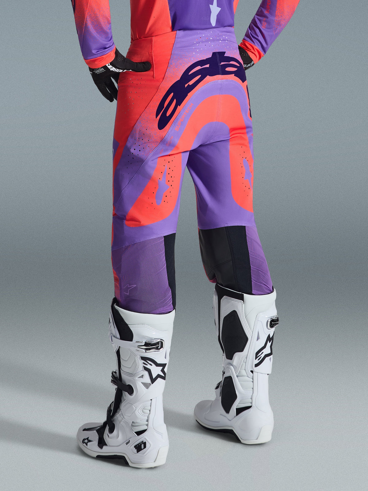 Alpinestars 2026 Supertech Scenz broek, motorcrossbroek, Hot Coral Purple, achteraanzicht met opvallende branding op het zitvlak, levendige koraalrode en paarse panelen met geperforeerde zones voor luchtstroom, gedragen met witte en zwarte motorcrosslaarzen