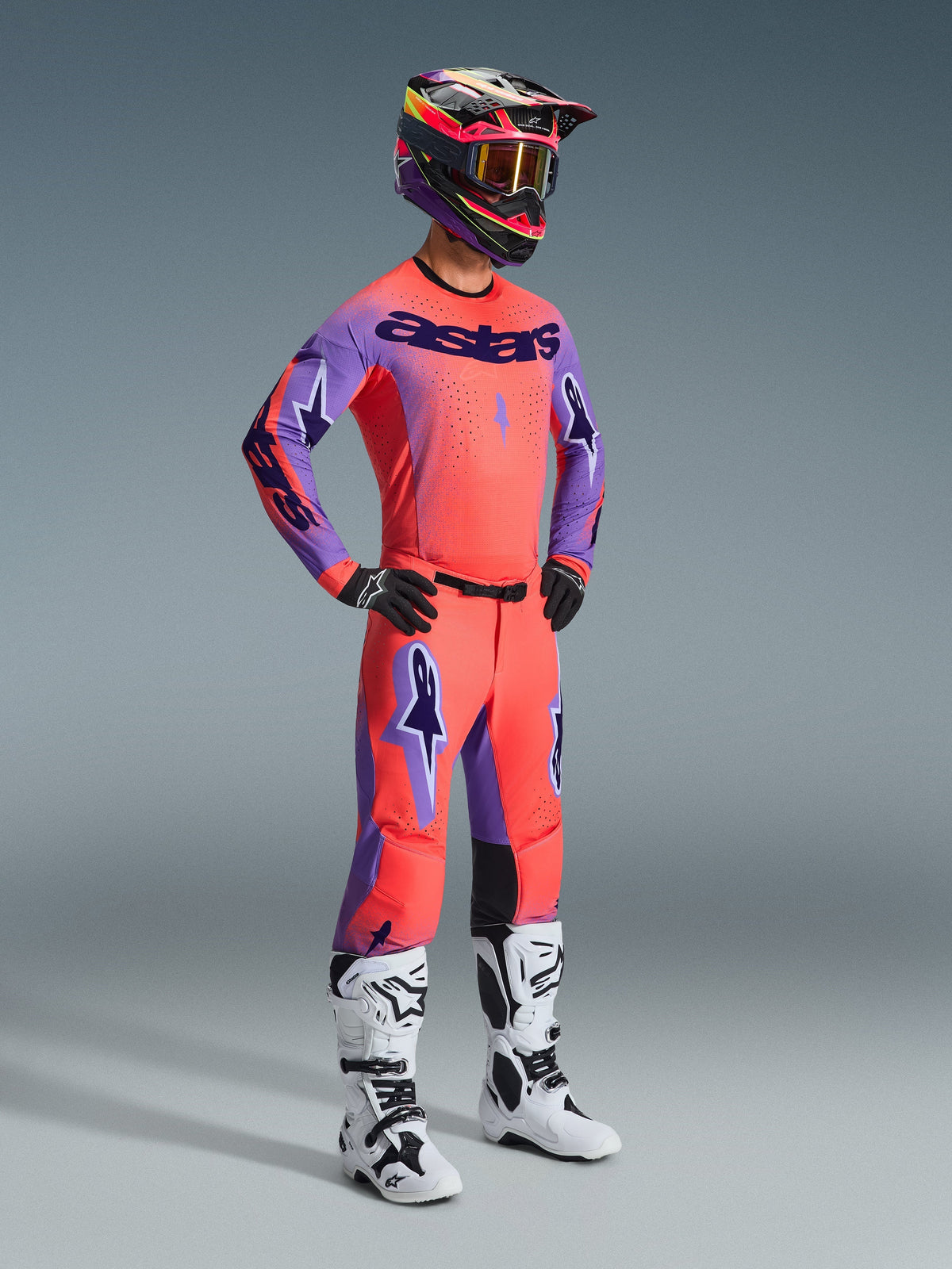 Motorcrosser in volledige race-uitrusting, voorzien van de Alpinestars 2026 Supertech Scenz Broek in Hot Coral Purple, bijpassend koraal en paars shirt, zwart-roze helm en wit-zwarte laarzen, professionele off-road racekit.
