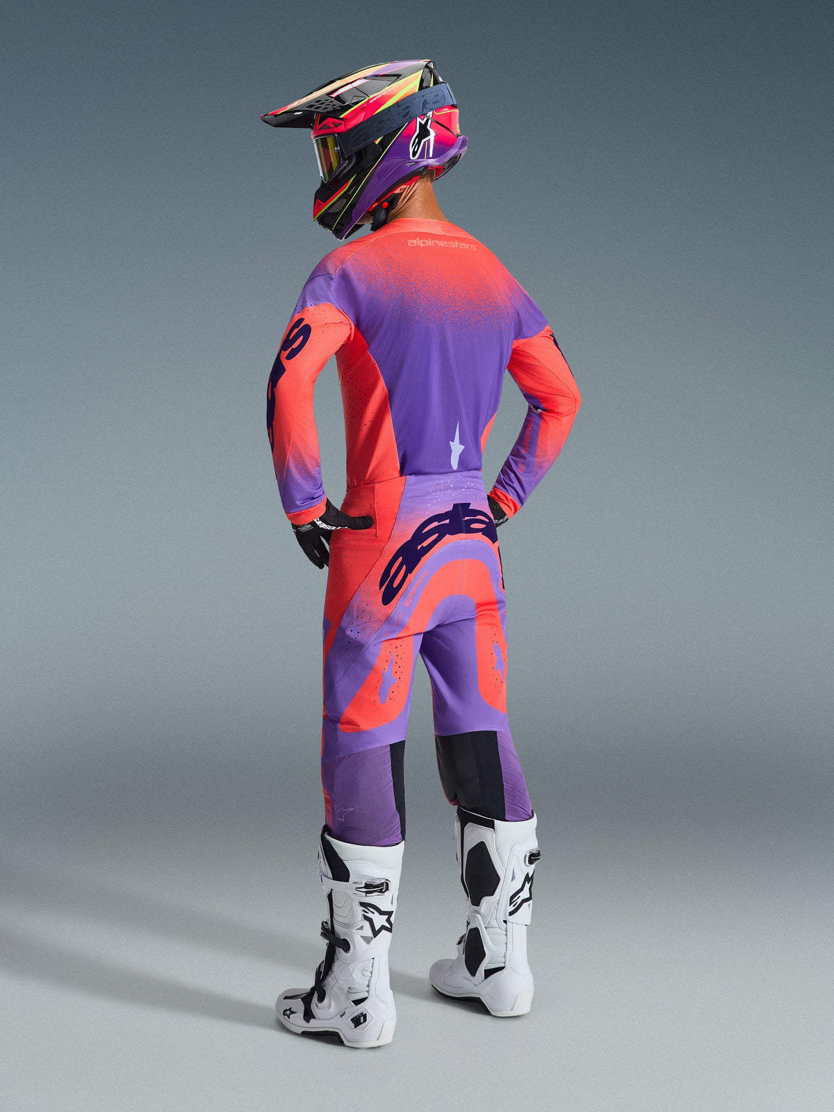 Alpinestars Supertech Scenz Broek 2026, motorcrossbroek, Hot Coral en paars, achteraanzicht met opvallende branding op het zitvlak, gedragen door een rijder met een bijpassend shirt en witte laarzen, professionele off-road race-uitrusting.
