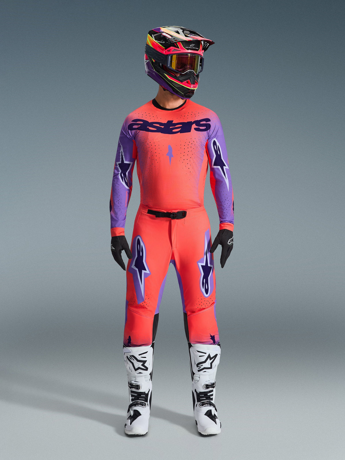 Alpinestars Supertech Scenz Broek 2026, motorcrossbroek, Hot Coral en paars, gedragen als een complete uitrusting met bijpassend shirt en helm, wit-zwarte laarzen, voorzien van paarse logo's en een atletische racepasvorm.