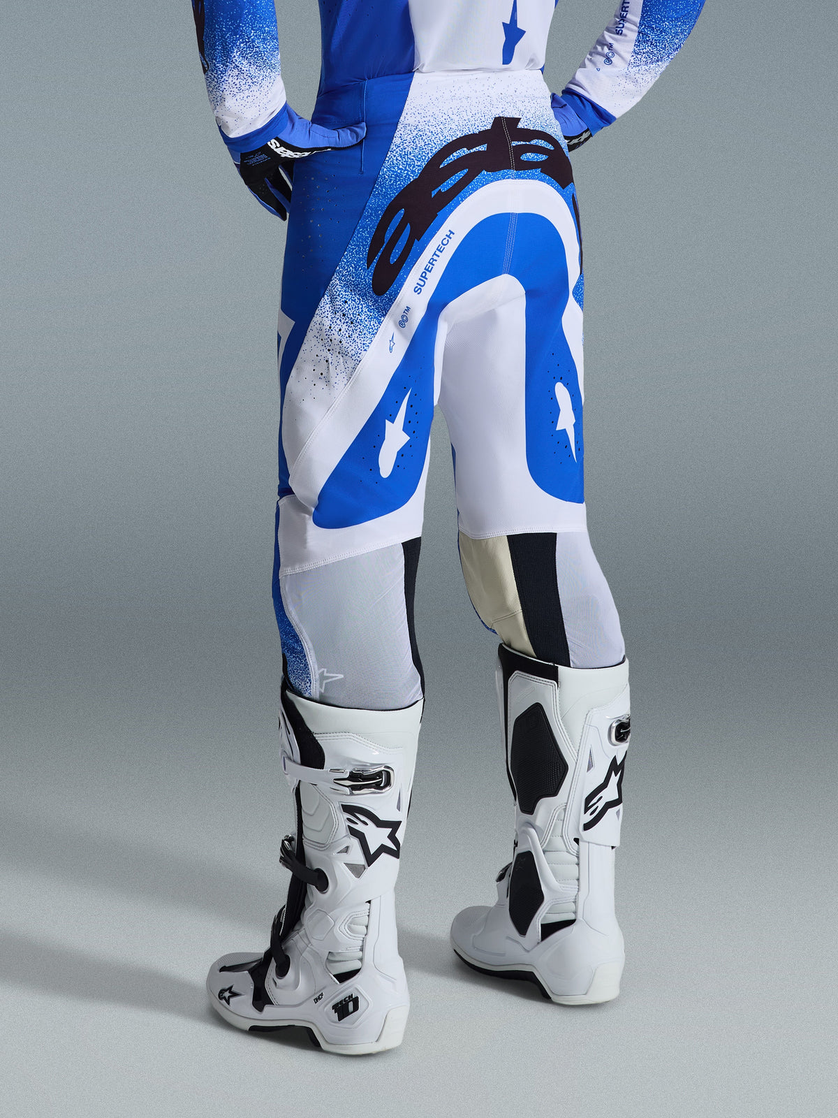 Pantalon Alpinestars Supertech Scenz 2026, pantalon de motocross, bleu Ucla et blanc, vue arrière montrant le grand logo Astars noir sur l'assise et le marquage 'SUPERTECH', porté avec des bottes Tech 10 blanches et un maillot bleu et blanc assorti.