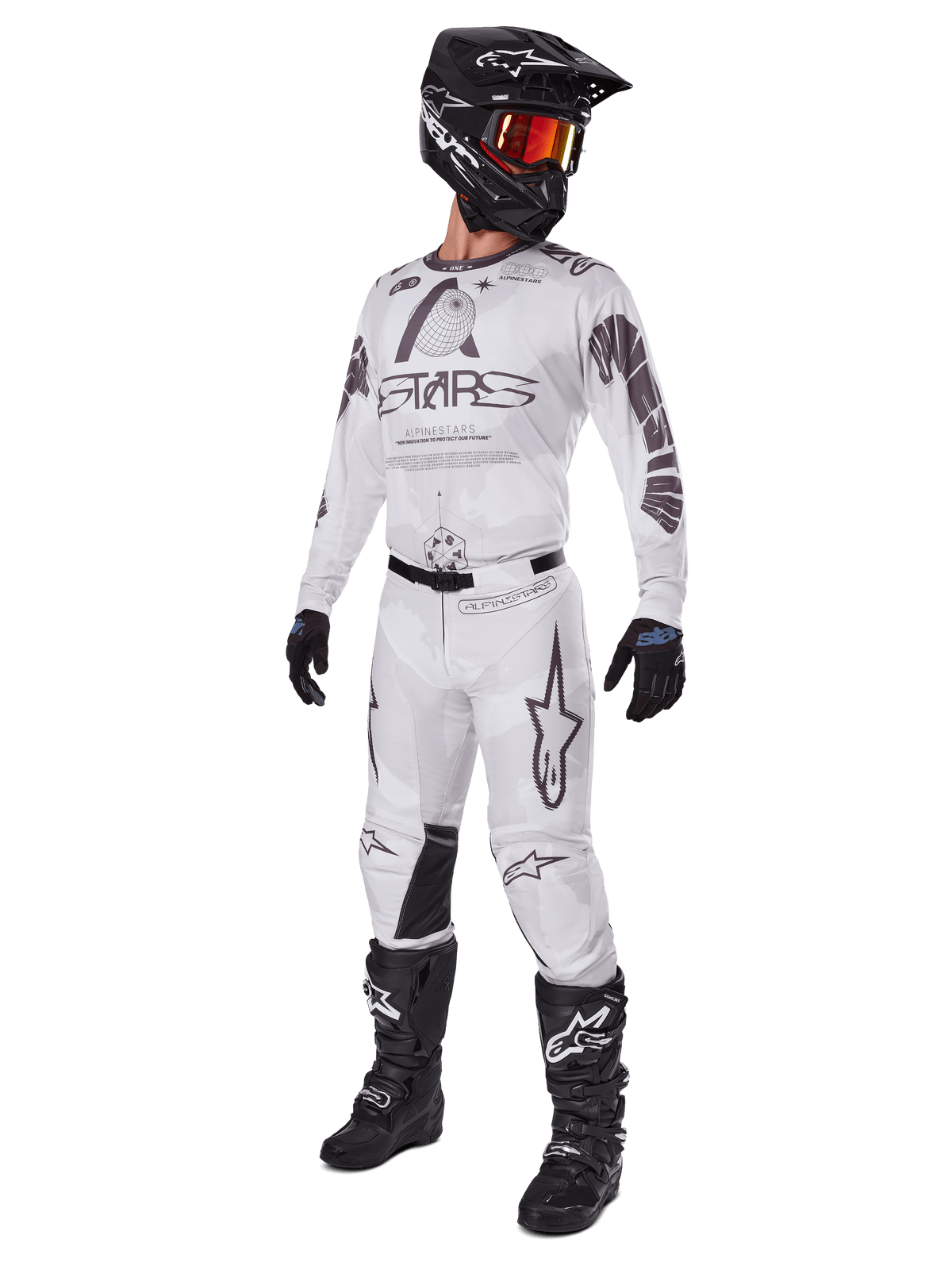 Alpinestars 2025 Racer Hollow broek, motorcross broek, lichtbruine camouflage, getoond als onderdeel van een complete race-uitrusting met een wit shirt, zwarte helm en zwarte laarzen, voorzien van versterkte zwarte kniepanelen en een subtiel camouflage-ontwerp.