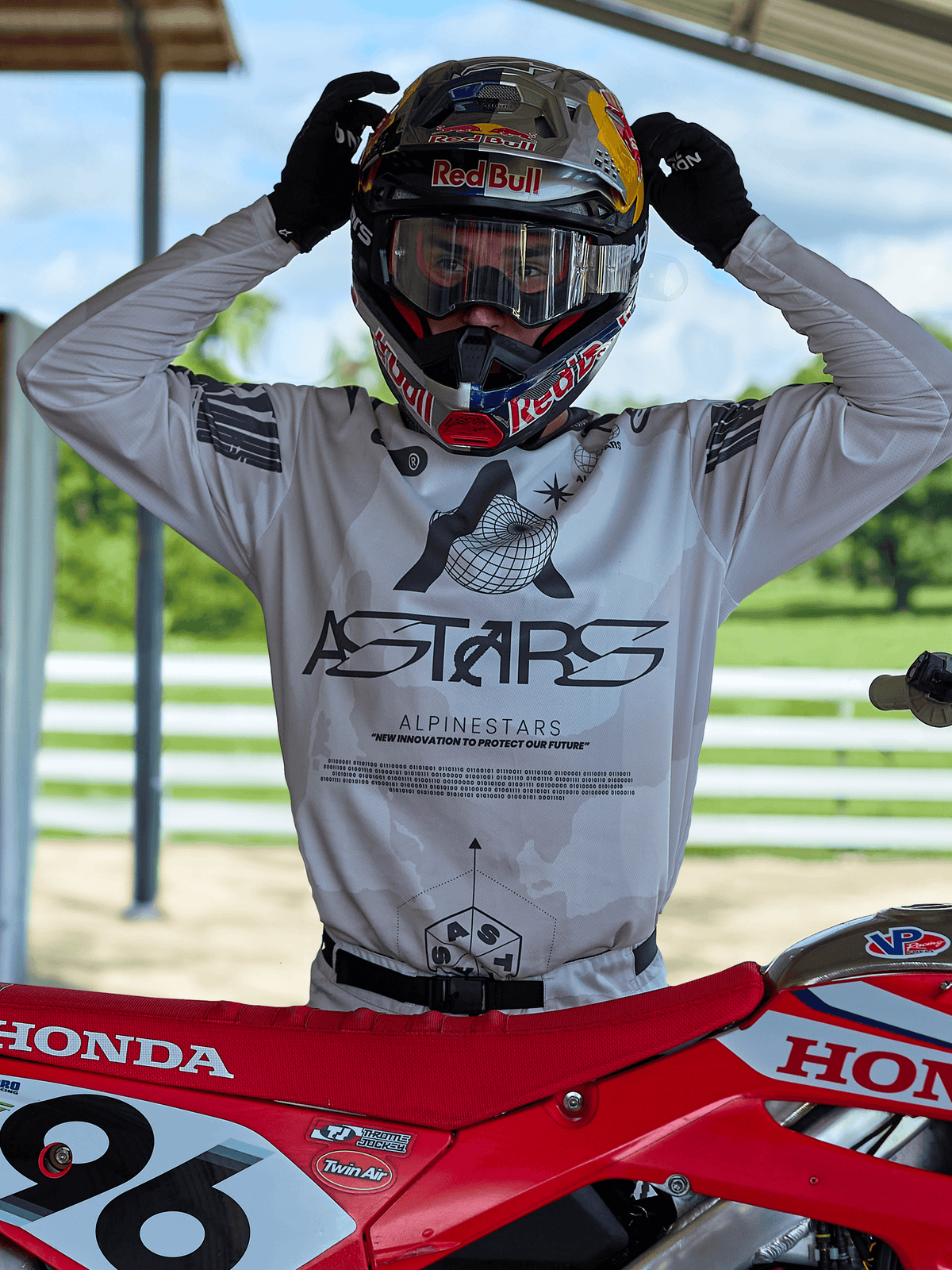 Motocrossrijder in Alpinestars 2025 Racer Hollow-uitrusting, lichte camo jersey met opvallende ASTARS-branding, stelt een Red Bull-helm met heldere crossbril af, staand naast een rode Honda crossmotor in een off-road omgeving.