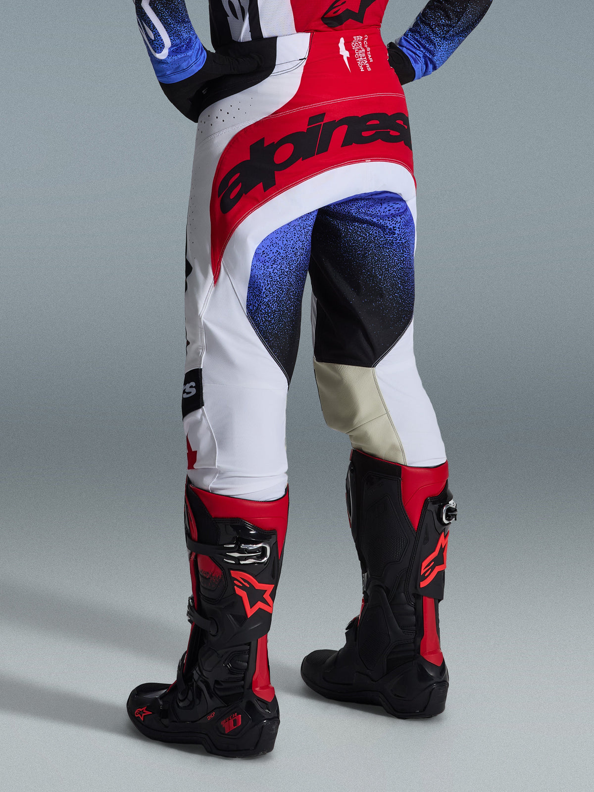 Alpinestars 2026 Techstar Knif Pants, pantalon de motocross, blanc, rouge et bleu, vue arrière montrant l'assise rouge avec logo noir, motifs mouchetés bleus et noirs sur les jambes, porté avec des bottes Tech 10 noires et rouges, équipement de course tout-terrain d'élite