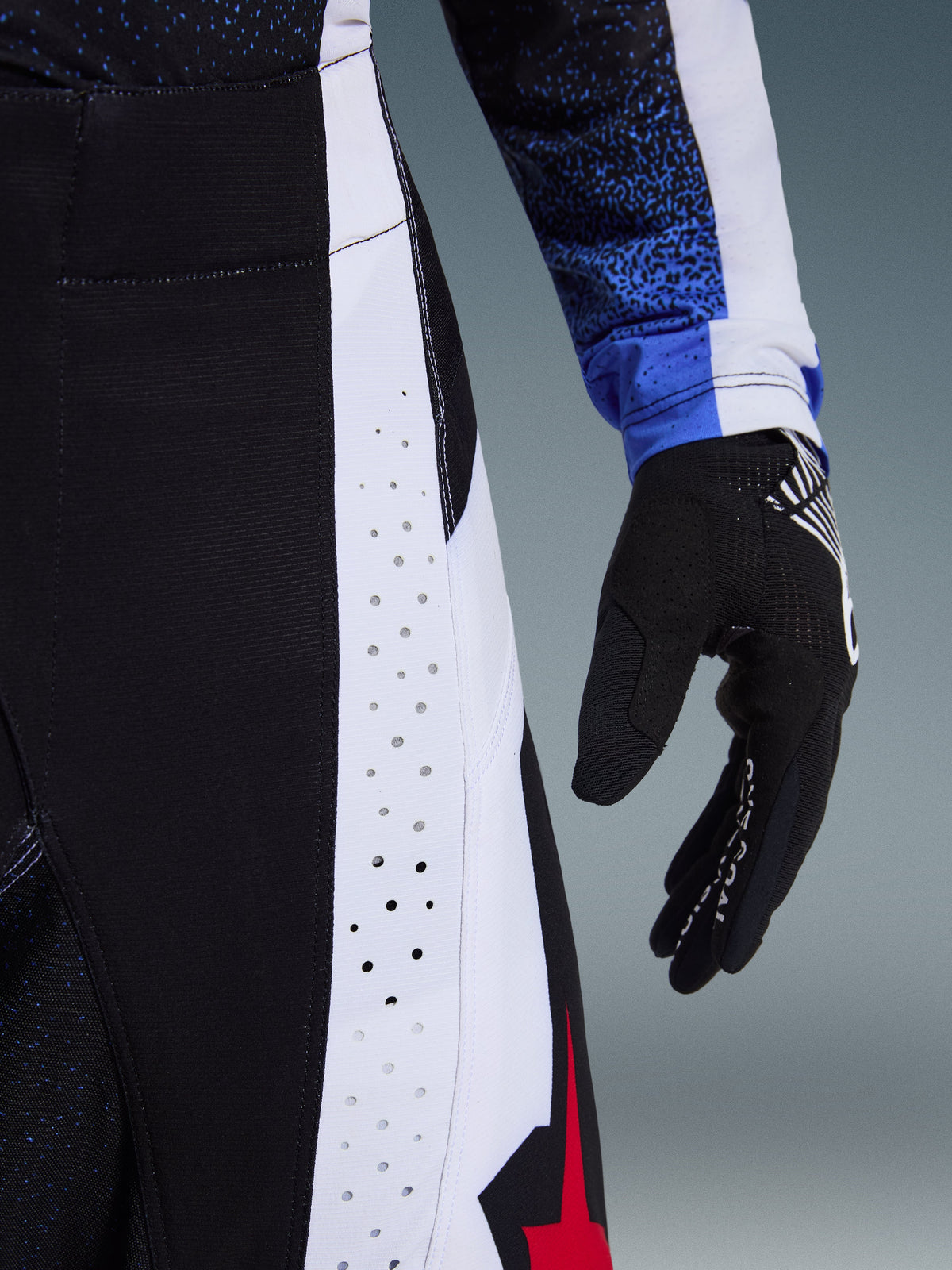 Alpinestars 2026 Techstar Knif Pants, pantalon de motocross noir, blanc et rouge, gros plan sur les perforations laser de ventilation, panneau latéral blanc, avec gant de motocross noir et maillot bleu à motifs, construction technique haute performance