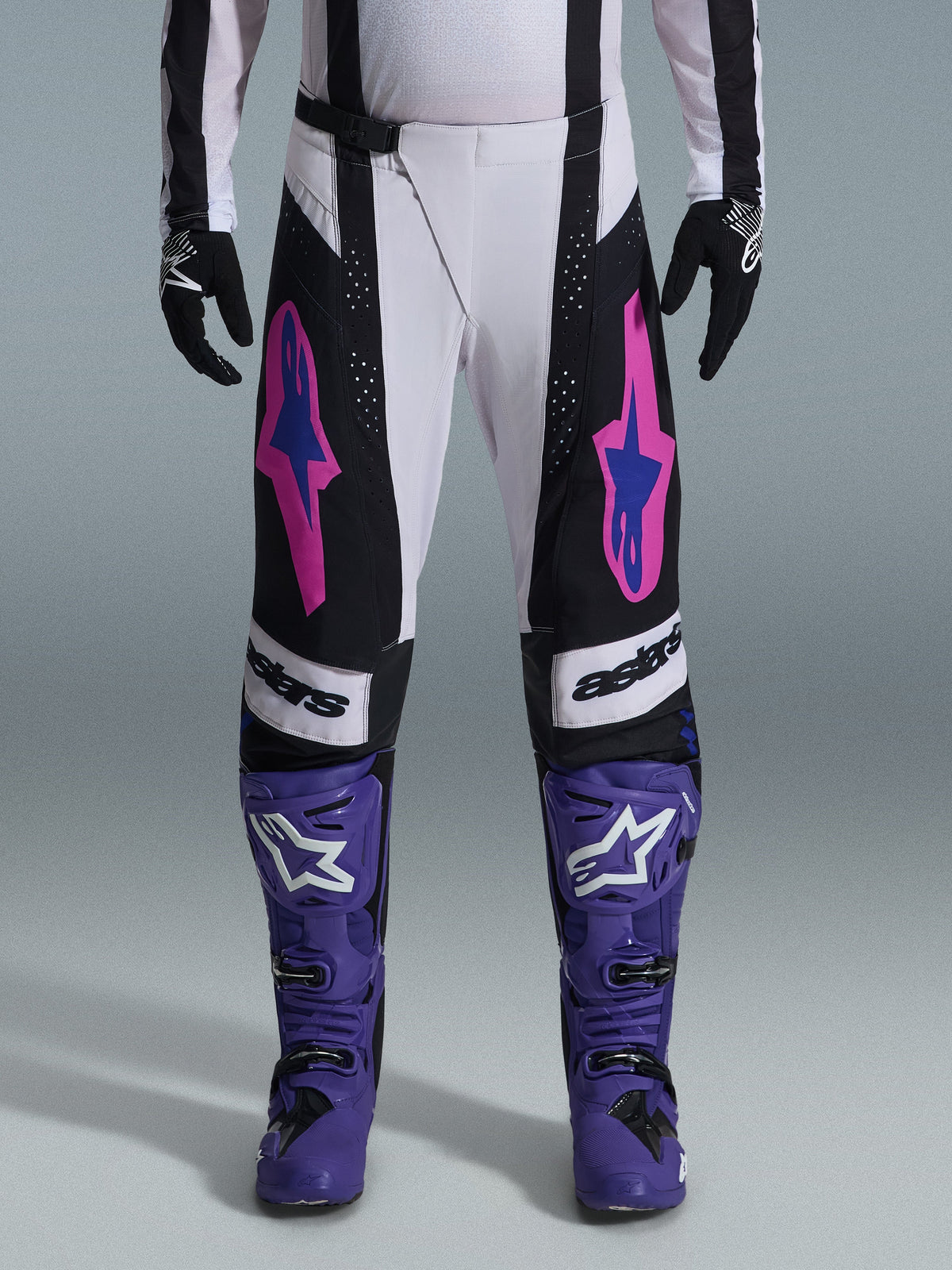 Alpinestars 2026 Techstar Knif Gear Set, Motocross-Fahrer in weiß-schwarzer Hose mit pinken und blauen Logos, lila Stiefel mit weißem Branding, schwarze Handschuhe, mit athletischer Rennpassform und lasergeschnittener Belüftung für den Offroad-Rennsport