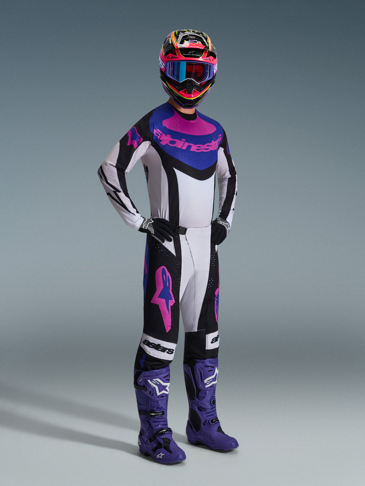 Motocross rider wearing the Alpinestars 2026 Techstar Knif Gear Set, Black Light Gray and Purple, white jersey with blue and pink accents, matching pants with purple star logos, colorful pink and yellow helmet, purple motocross boots, and black gloves, professional gear set for off-road racing.