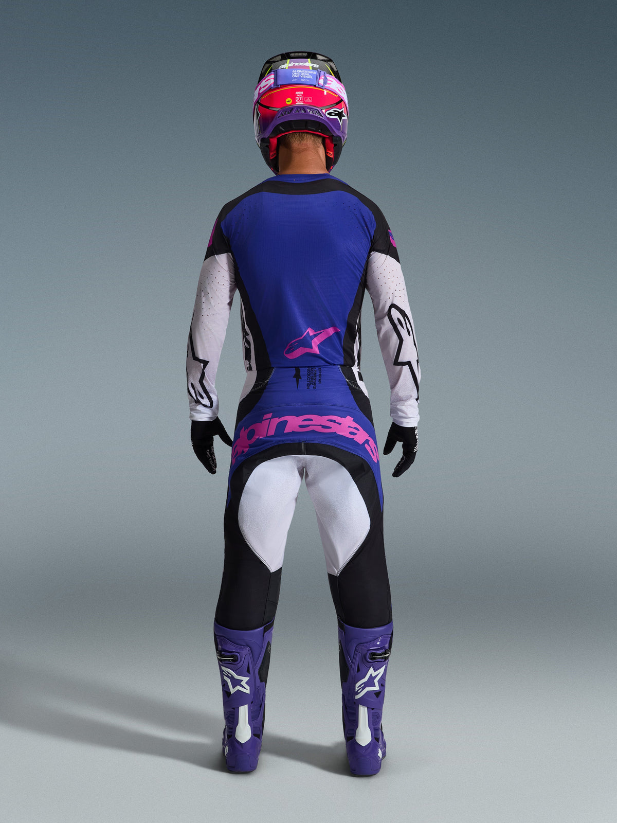 Rückansicht eines Motocross-Fahrers im Alpinestars 2026 Techstar Knif Gear Set, blau-weißes Trikot mit pinkem Stern-Logo, dazu passende blau-schwarze Hose mit großem pinkem Alpinestars-Branding auf der Rückseite, lila-weiße Stiefel und lila-roter Helm, professionell abgestimmtes Offroad-Rennoutfit