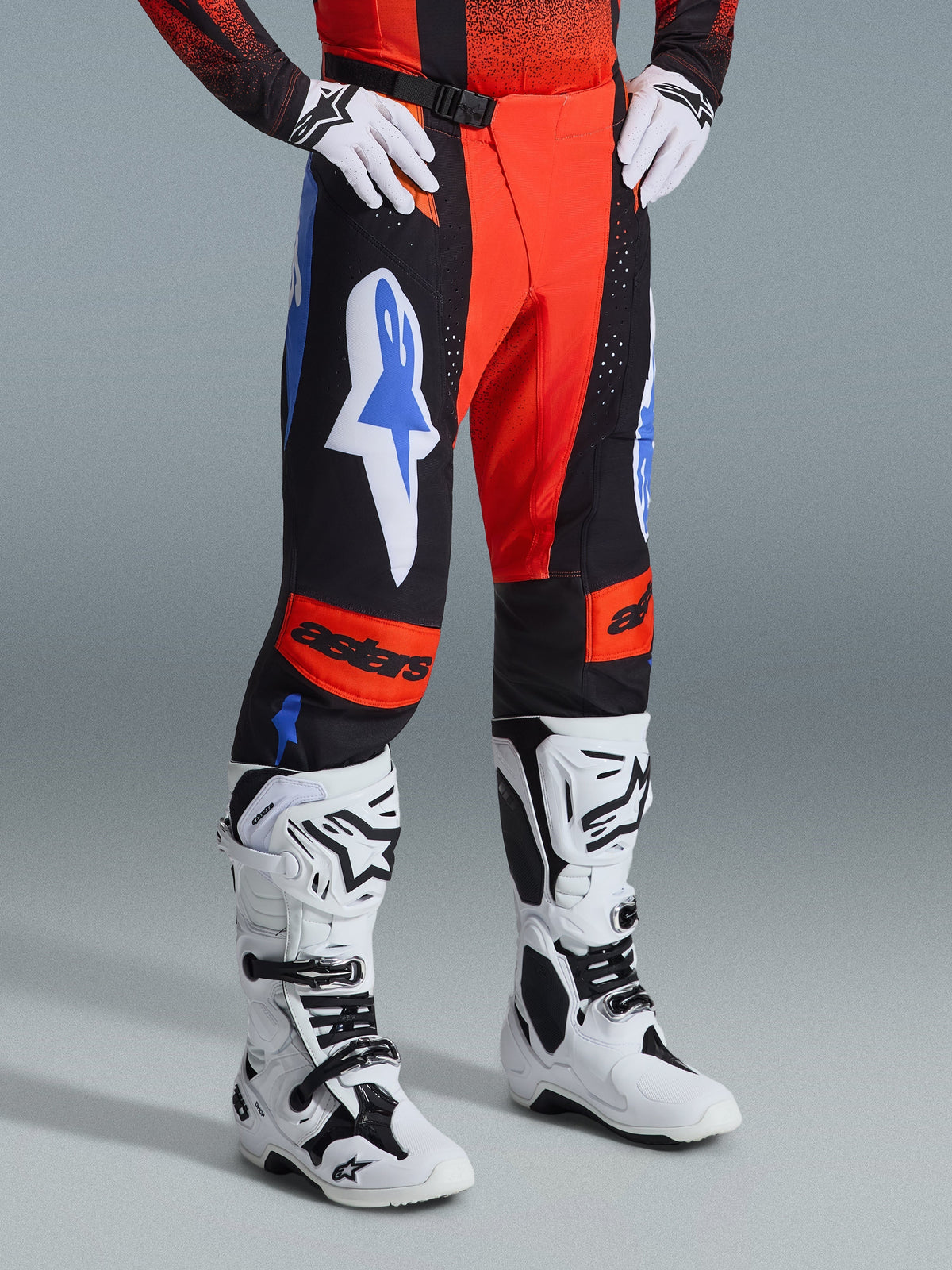 Alpinestars Techstar Knif Broek 2026, motorcrossbroek, oranje, zwart en UCLA blauw, gedragen met witte handschoenen en wit-zwarte motorcrosslaarzen, met opvallende logo's op de dijen en oranje kniepanelen met astars-branding.