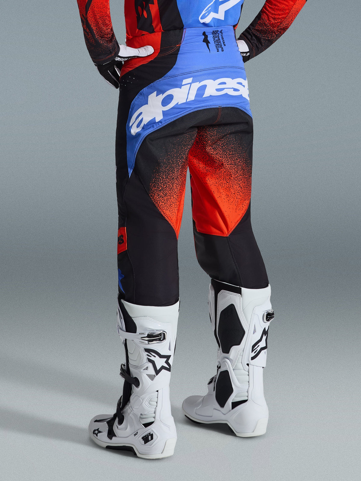 Alpinestars 2026 Techstar Knif crossbroek, motorcrossbroek, oranje, zwart en UCLA-blauw, achteraanzicht met blauw achterpand en wit Alpinestars-logo, oranje kleurverloop op de pijpen, gedragen met bijpassende jersey en witte laarzen voor off-road racen
