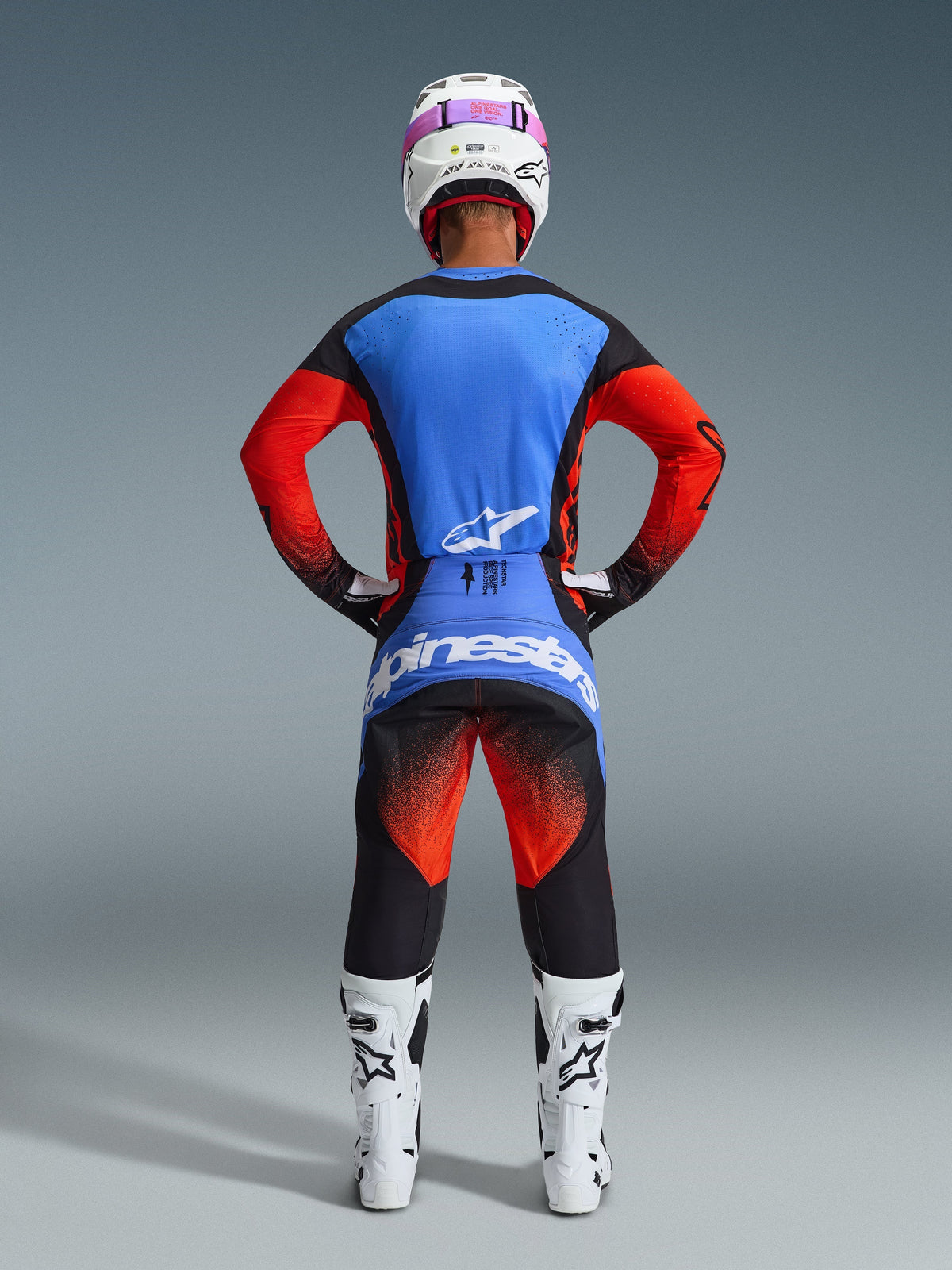 Motocrossrijder van achteren gezien in de 2026 Techstar Knif Broek in oranje, zwart en UCLA blauw, gecombineerd met een blauw en oranje jersey, witte helm met roze bril en witte racelaarzen, volledige Alpinestars off-road competitie-uitrusting