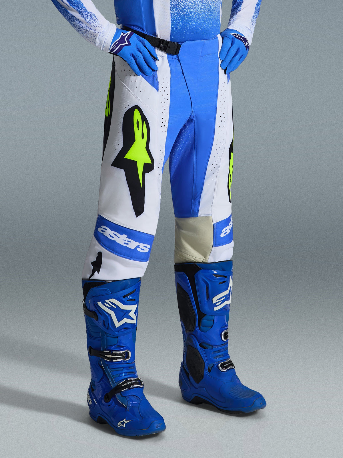 Alpinestars Techstar Knif Pants 2026, pantalon de motocross, bleu UCLA, noir et jaune fluo, porté avec des bottes et des gants de motocross bleus assortis, avec des panneaux blancs, de grands logos étoilés jaune fluo et un design athlétique tout-terrain
