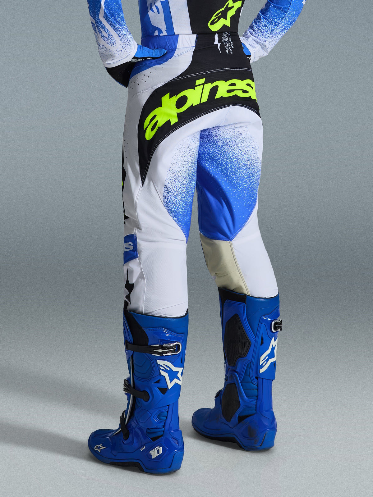 Pantalon Alpinestars Techstar Knif 2026, pantalon de motocross, bleu Ucla, noir et jaune fluo, vue arrière montrant un logo Alpinestars jaune fluo audacieux dans le dos, châssis blanc avec graphismes en dégradé bleu, porté avec des bottes de motocross bleu vif haute performance