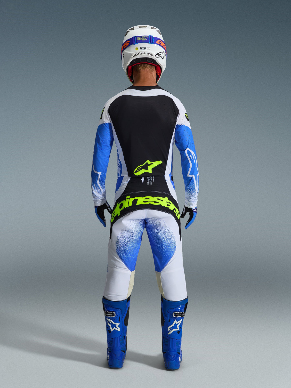 Pilote de motocross vu de dos, tenue de course complète, pantalon Alpinestars Techstar Knif 2026 bleu UCLA, noir et jaune fluo, maillot à manches longues assorti avec empiècements noirs et blancs, bottes tout-terrain bleues, casque blanc, logo Alpinestars jaune fluo sur l'assise
