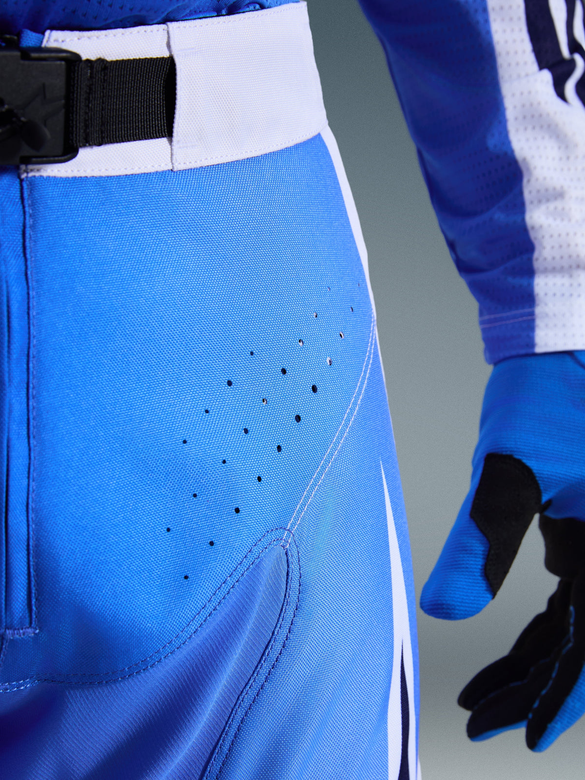 Pantalon Alpinestars Techstar Knif 2026, bleu UCLA et blanc, gros plan sur la zone de la hanche montrant les perforations découpées au laser pour la ventilation, fermeture à la taille par boucle magnétique SX2 et gants de motocross assortis
