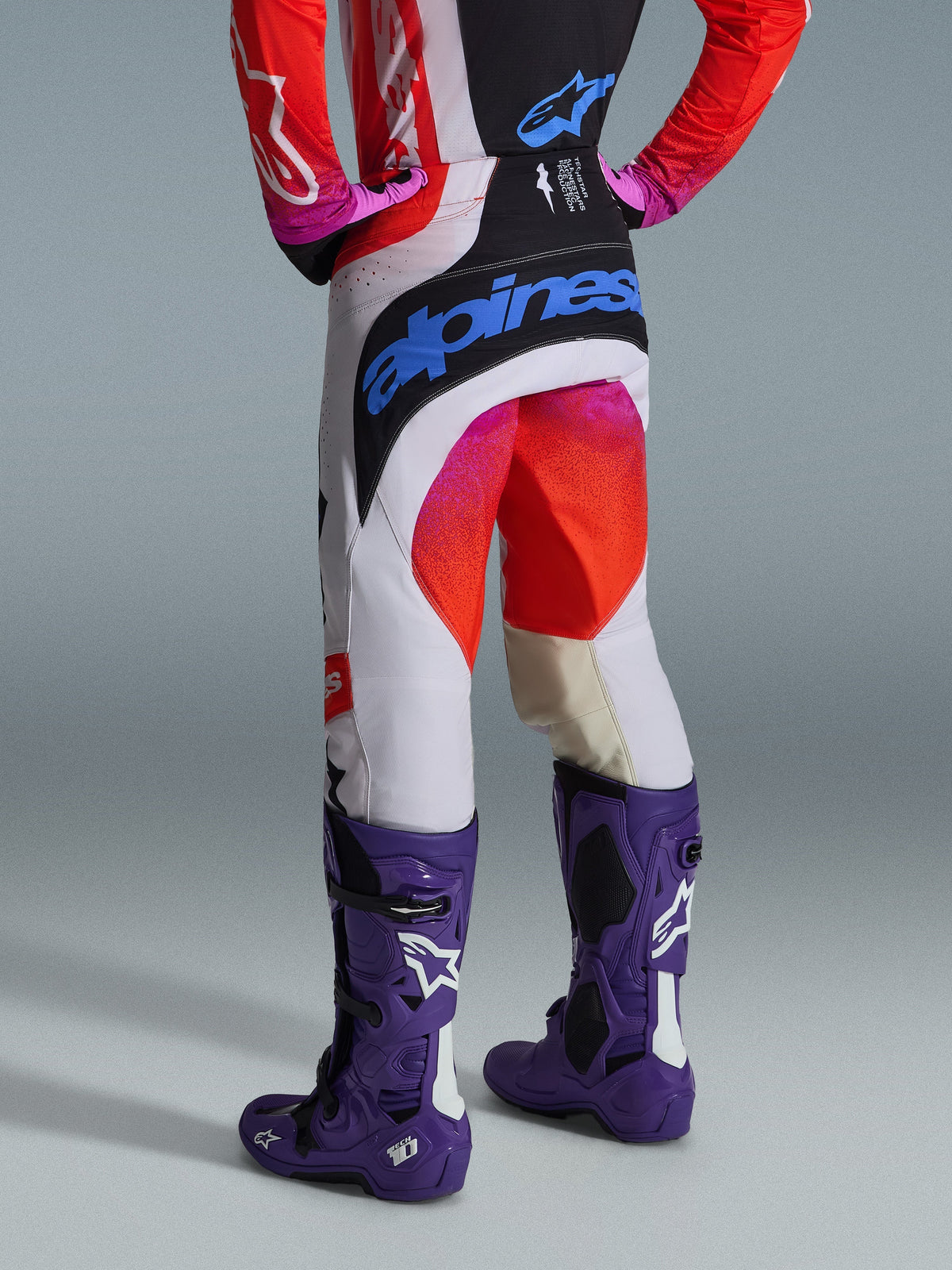 Alpinestars 2026 Techstar Knif broek en kledingset, achteraanzicht van een motorcrosser met een witte broek met zwarte panelen, roze en rode kleurverloop-accenten en een groot blauw Alpinestars-logo op het zitvlak, gecombineerd met paarse Tech 10-laarzen en een bijpassende rode en zwarte jersey.