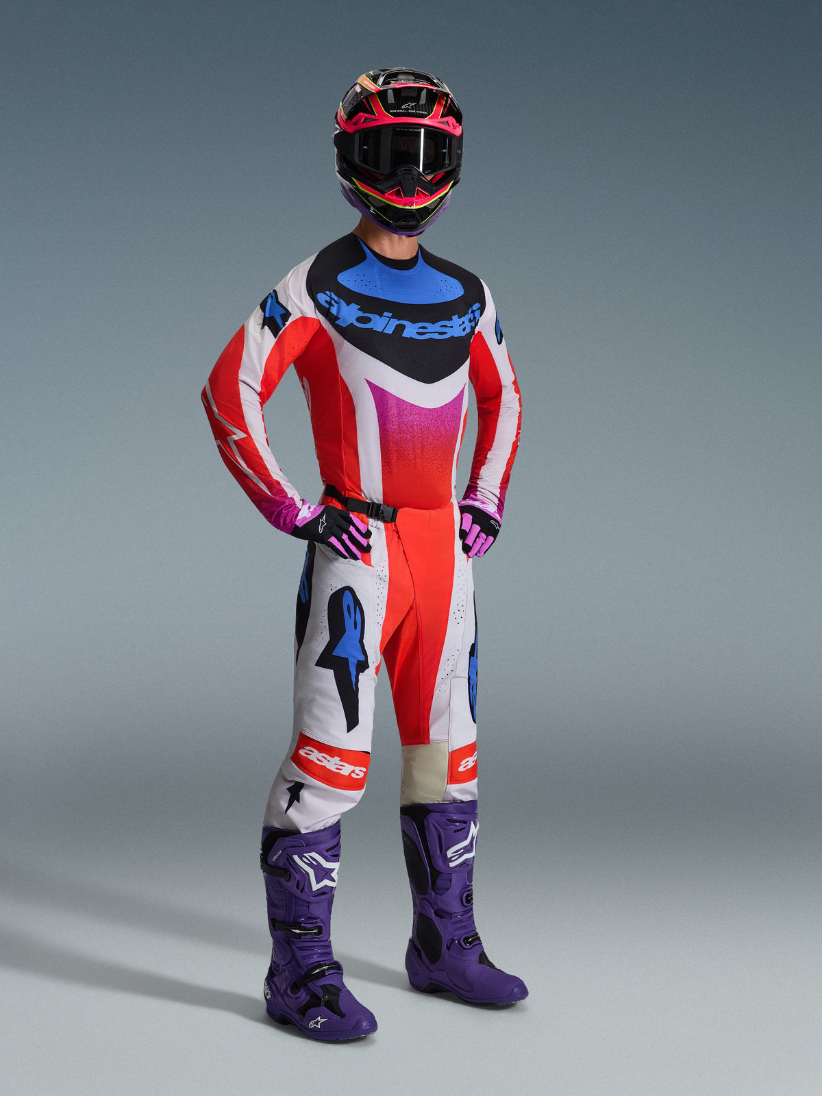 Pilote de motocross portant un équipement de course complet Alpinestars, maillot Techstar Knif 2026 blanc et rouge avec des accents bleus et roses, pantalon de motocross assorti, bottes violettes et casque multicolore, tenue professionnelle coordonnée pour la performance tout-terrain