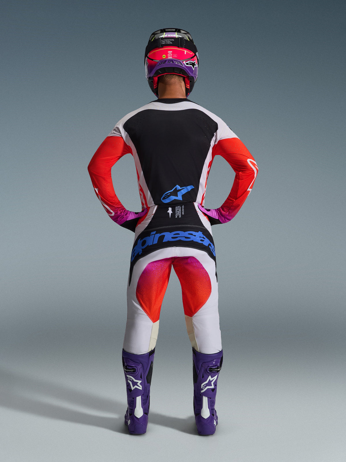 Tenue de motocross complète Alpinestars Techstar Knif 2026 vue de dos, maillot gris clair avec panneau arrière noir et manches orange, pantalon blanc et orange avec logo bleu, bottes violettes et casque noir et rose, équipement professionnel pour le tout-terrain