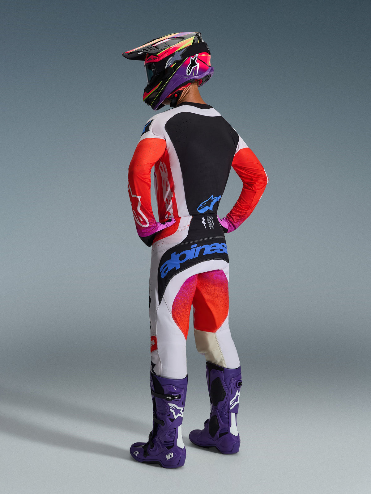 Vue arrière d'un pilote de motocross portant le maillot Alpinestars 2026 Techstar Knif, gris clair et noir avec des manches orange fluo, associé à un pantalon de course assorti, des bottes violettes et un casque multicolore, équipement de motocross professionnel.